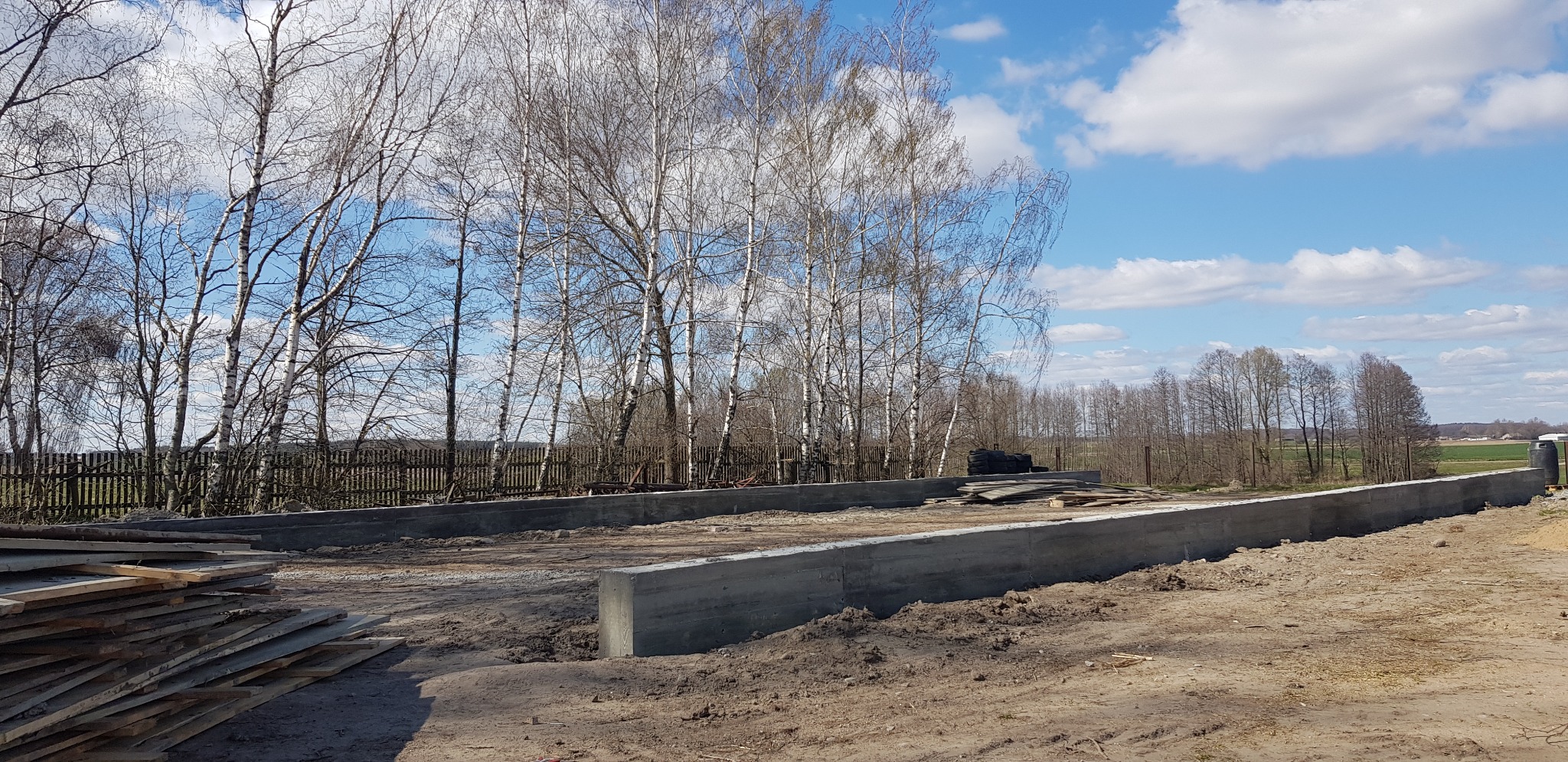 Wylane betonowe fundamenty pod budowę domu na działce z widocznymi drzewami i niebem z chmurami.