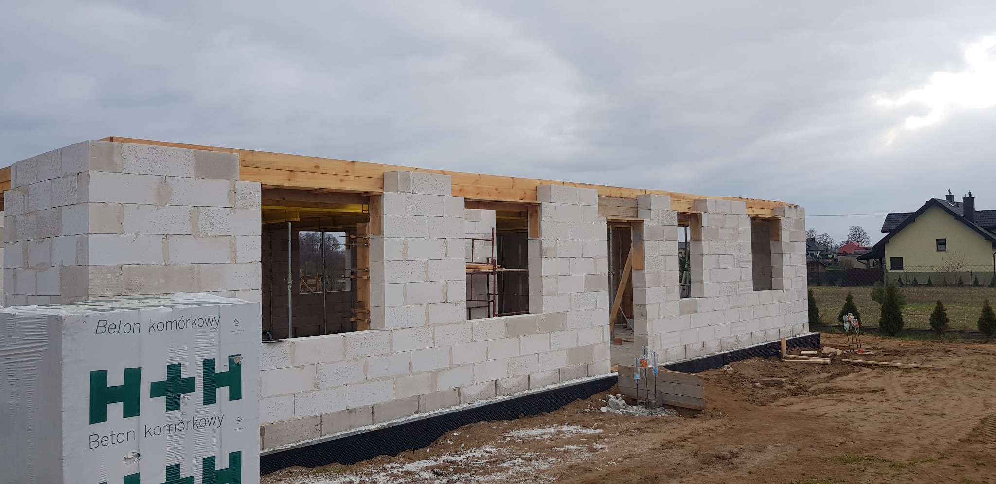 Wznoszenie ścian parterowego budynku z betonu komórkowego H+H, widoczne drewniane nadproża okienne i paleta z bloczkami na pierwszym planie.