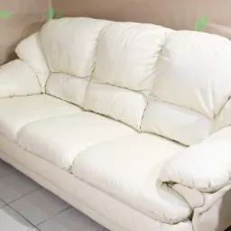 Jasna, skórzana sofa trzyosobowa z miękkimi poduchami na siedzisku i oparciu, częściowo osłonięta papierem malarskim zabezpieczającym ścianę.