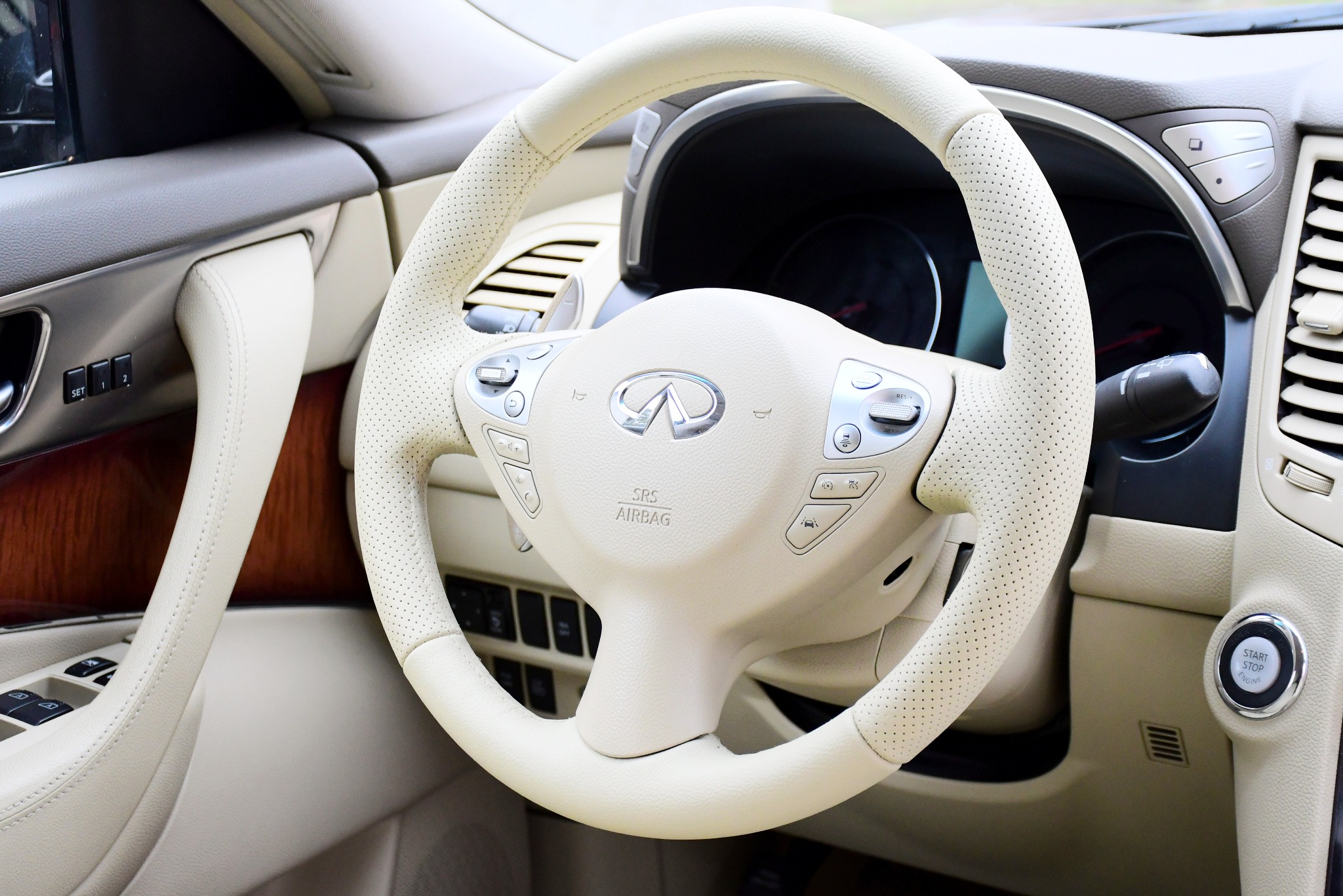 Beżowa, perforowana skórzana kierownica w samochodzie marki Infiniti, widoczna deska rozdzielcza i przycisk start/stop silnika. Wnętrze pojazdu w Nasielsku po renowacji.