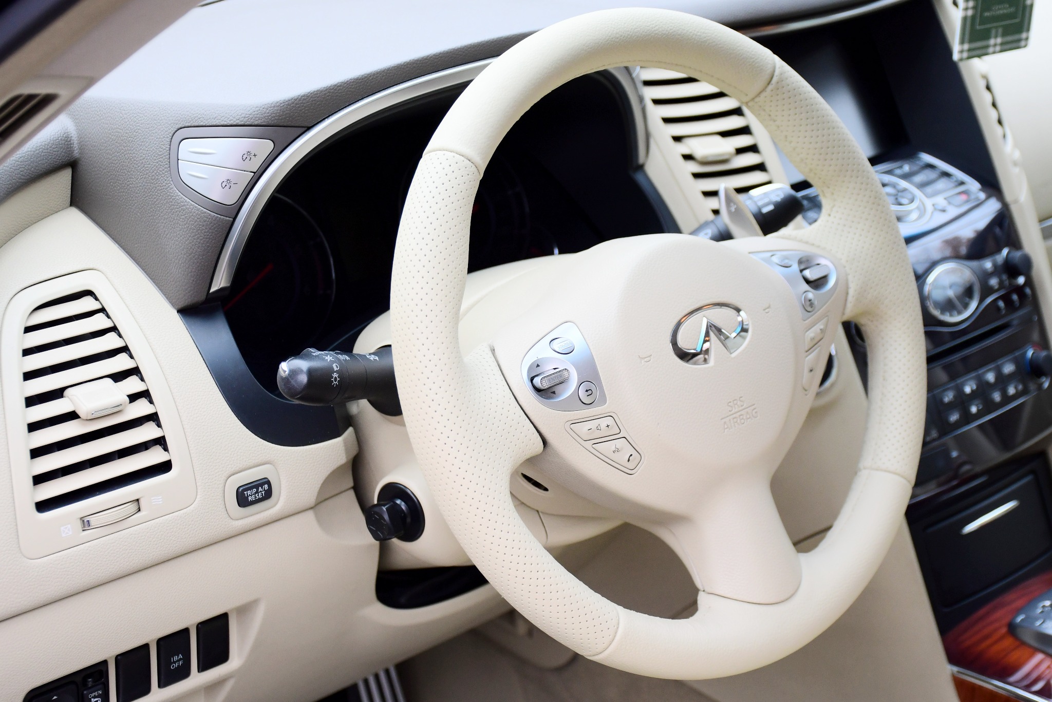 Beżowa, perforowana skórzana kierownica z logo Infiniti w luksusowym samochodzie, widok na deskę rozdzielczą i przyciski sterowania.