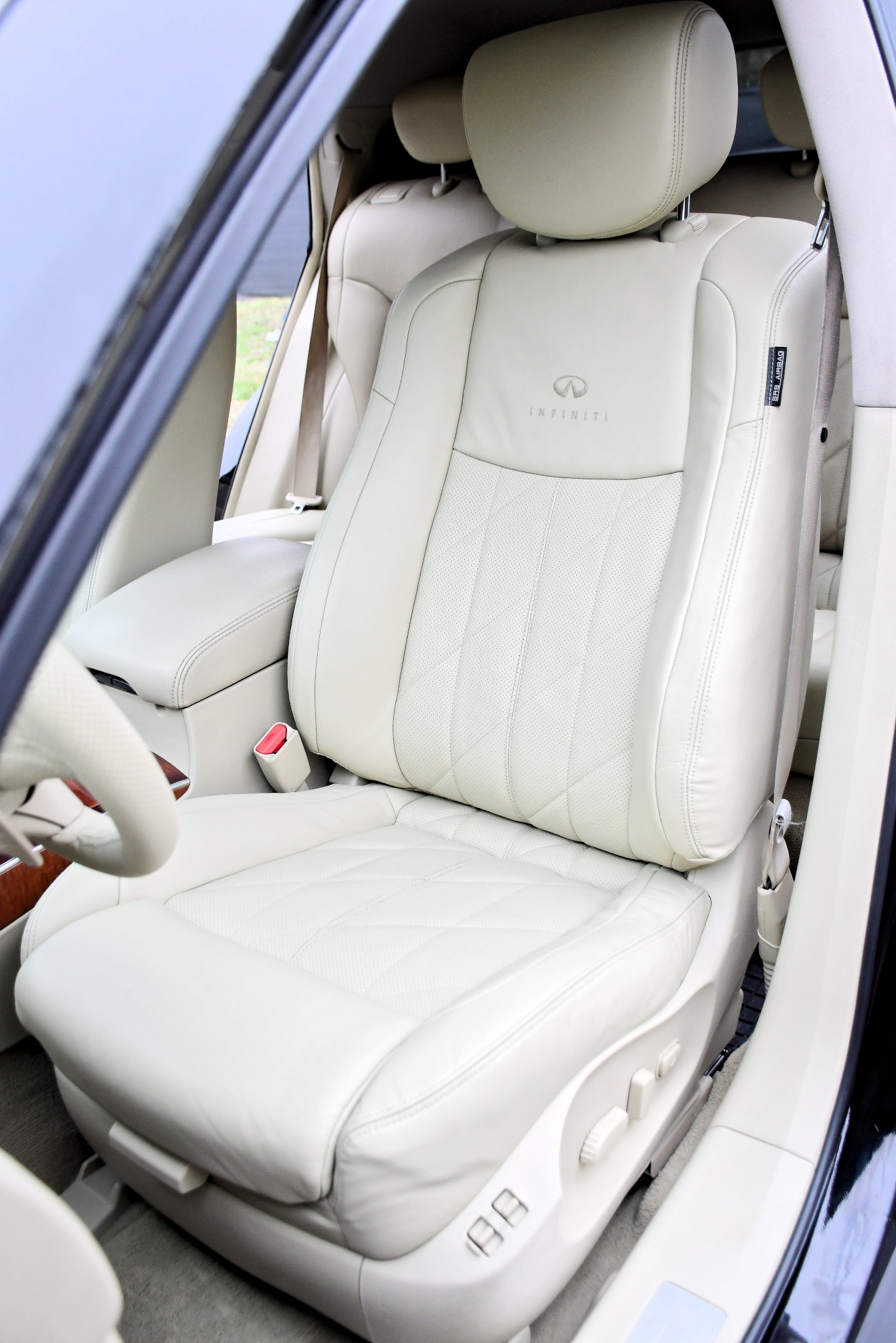 Jasny, skórzany fotel samochodowy z wytłoczonym logo Infinity na zagłówku, widoczne elementy regulacji i pas bezpieczeństwa.