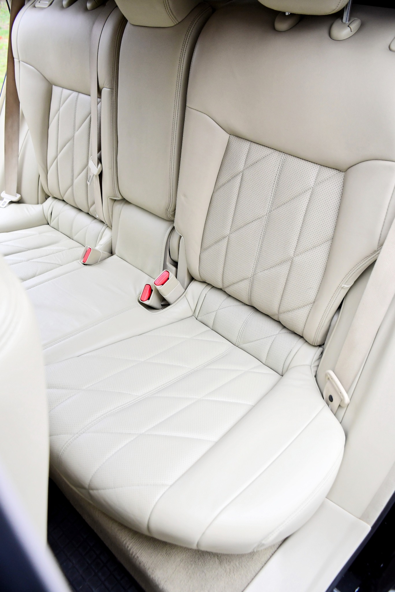 Jasna, skórzana tylna kanapa samochodu z pikowaniem w romby i czerwonymi elementami pasów bezpieczeństwa.