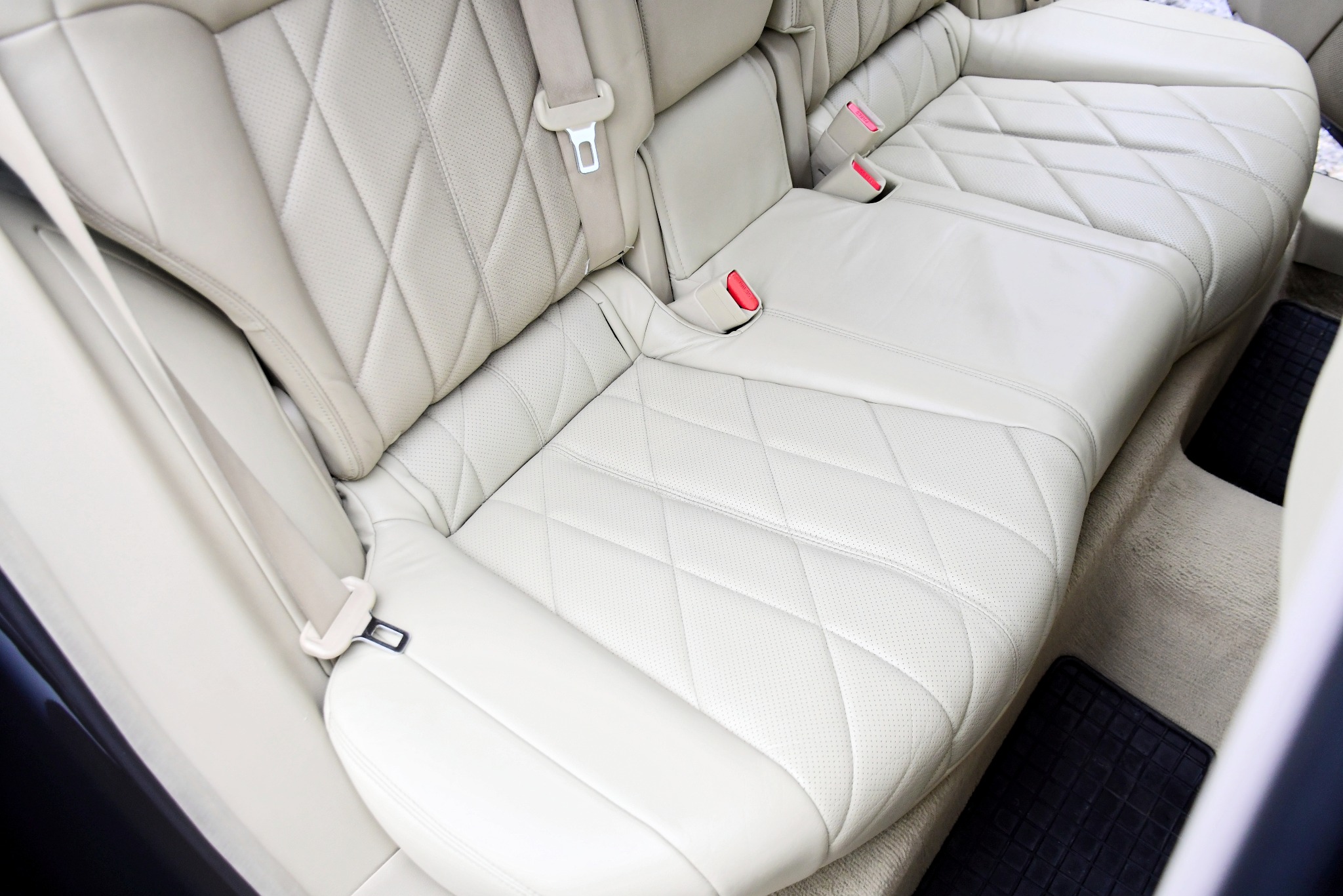 Jasna, skórzana tylna kanapa samochodowa z pikowaniem w romby i widocznymi pasami bezpieczeństwa oraz czarnymi dywanikami podłogowymi.