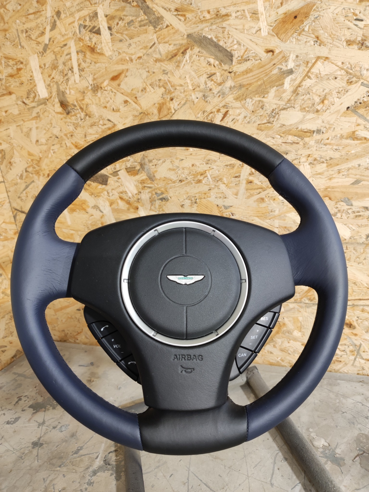 Odnowiona, dwukolorowa kierownica samochodowa z logo na tle drewnianej płyty. Górna część obszyta czarną skórą, boki w kolorze niebieskim, widoczne przyciski sterowania i napis AIRBAG.
