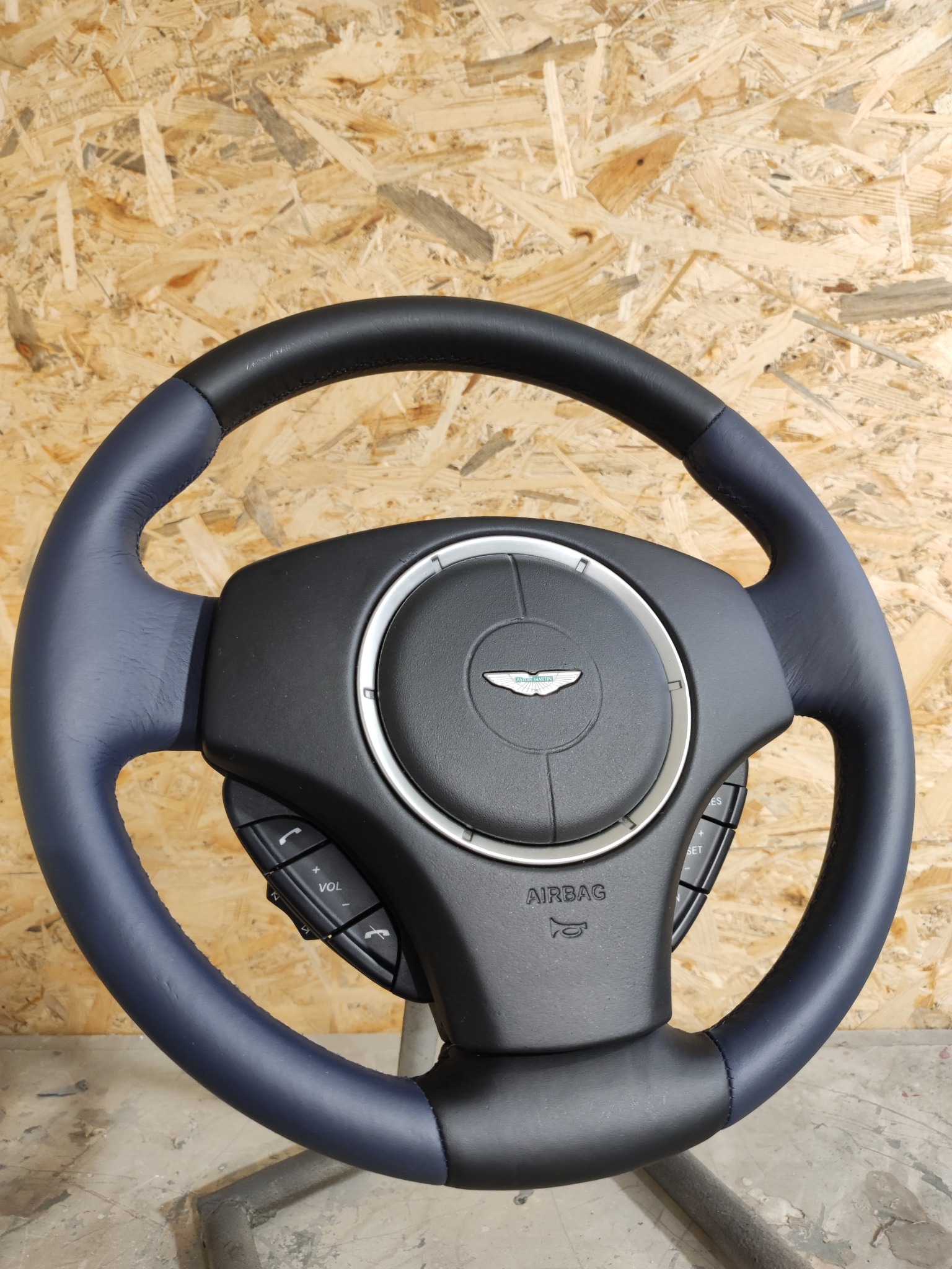 Odnowiona kierownica samochodowa Aston Martin, obszyta skórą w kolorze granatowym i czarnym, widoczne przyciski sterowania i logo marki na tle drewnianej ściany.