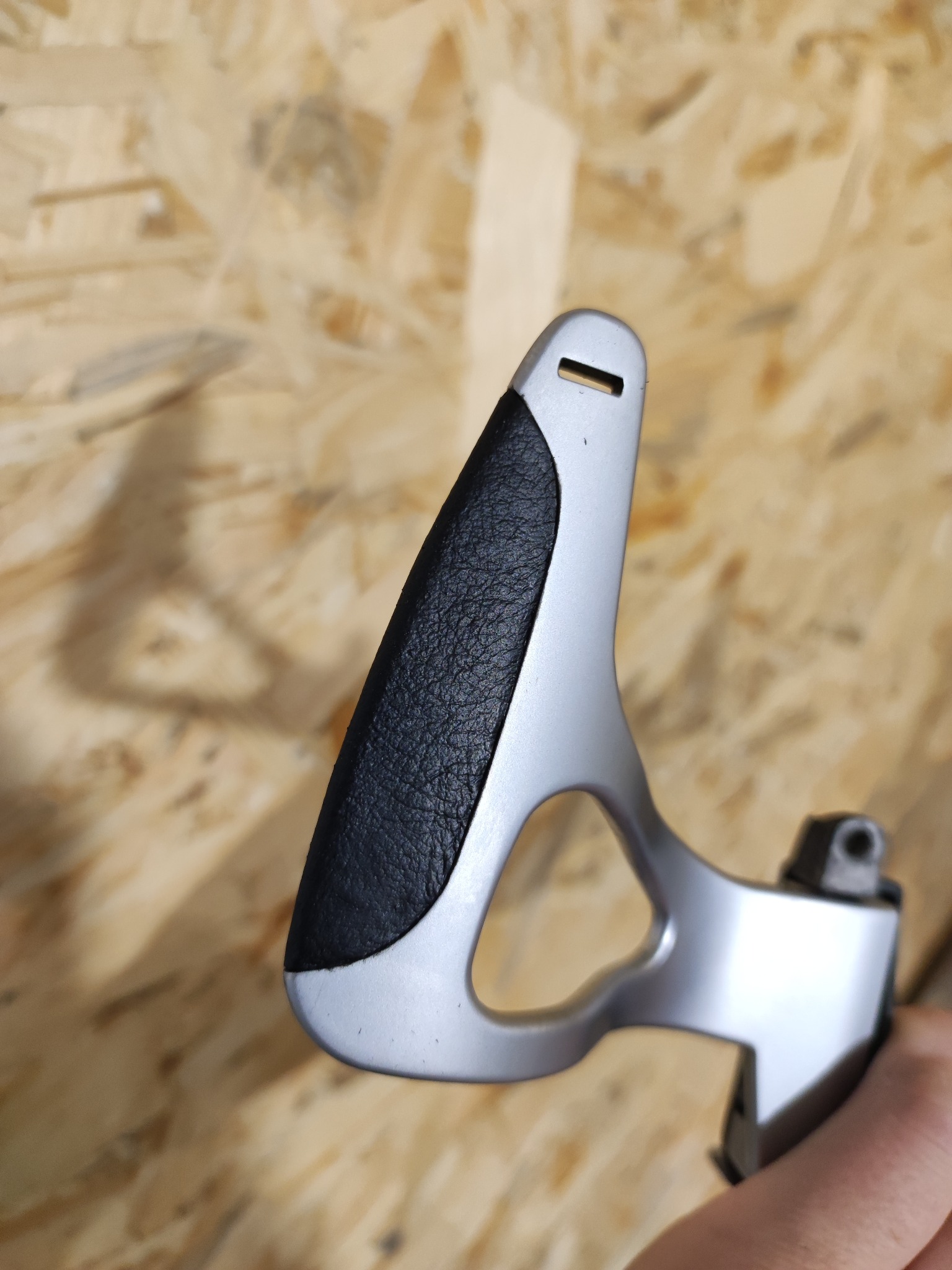 Metalowa rączka dźwigni zmiany biegów z czarnym skórzanym uchwytem na tle płyty OSB.