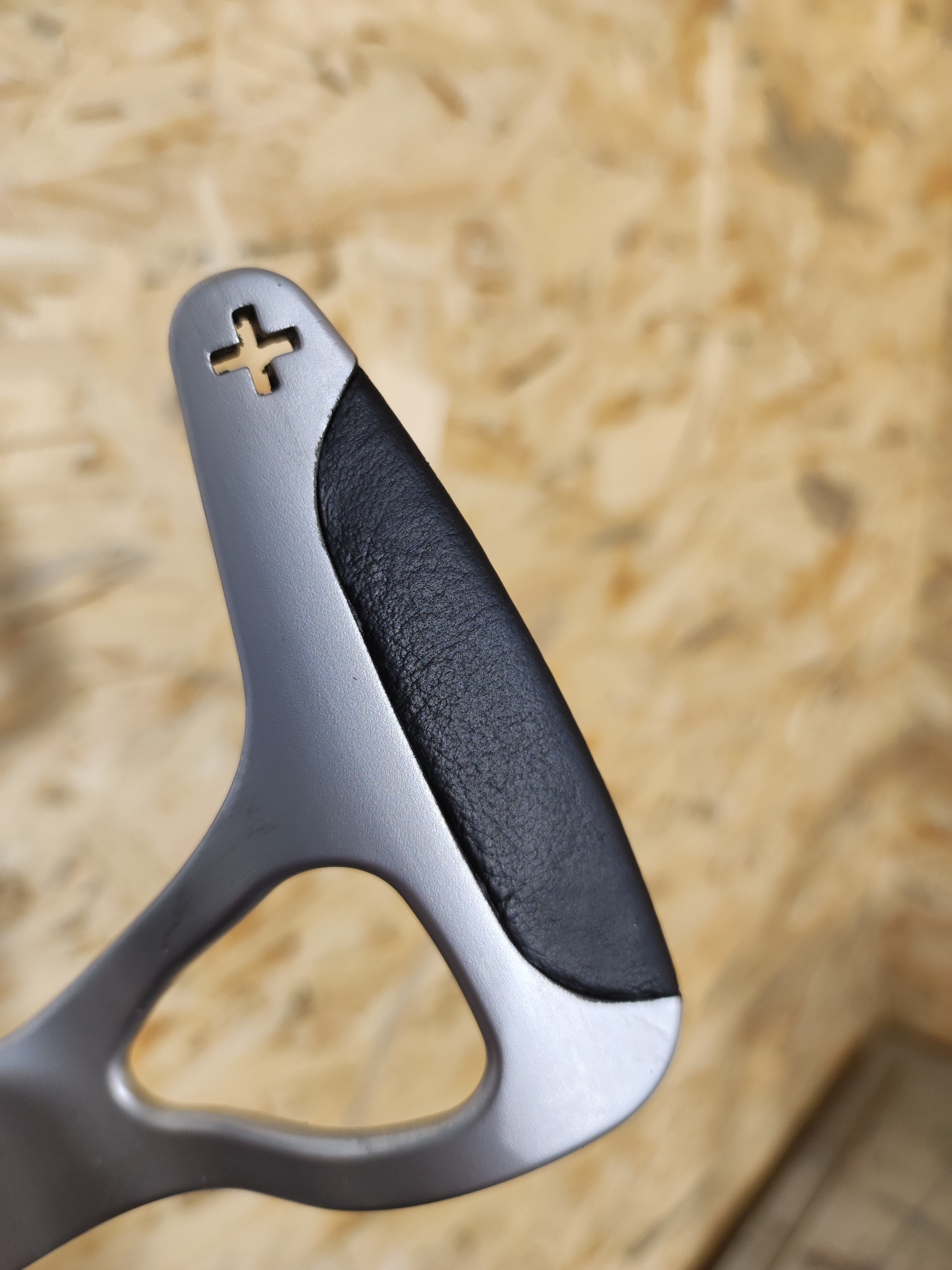 Szczegół dźwigni zmiany biegów z elementem obszytym czarną skórą i wyciętym znakiem plusa, na tle płyty OSB.