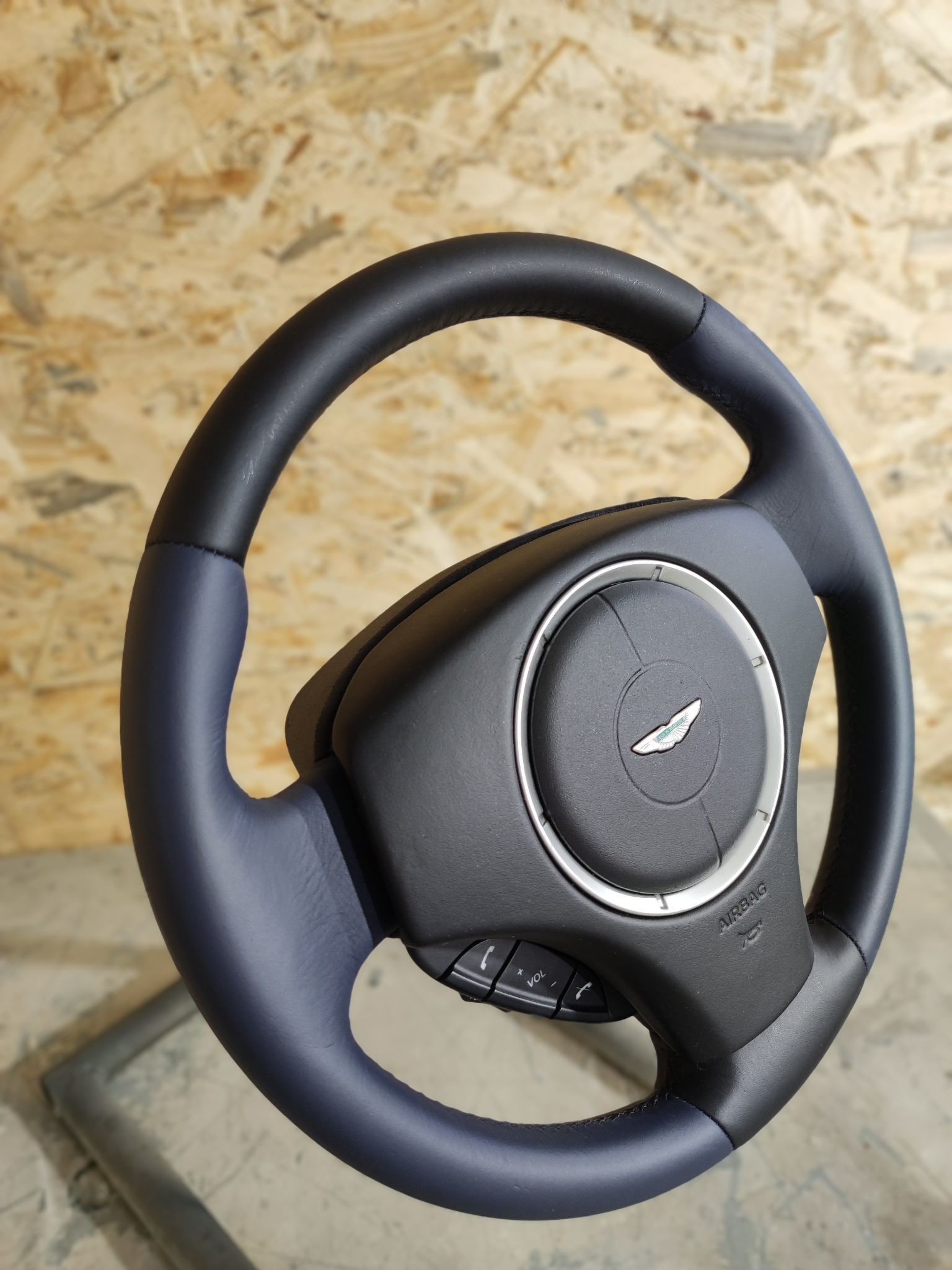 Odnowiona kierownica samochodowa, obszyta skórą w kolorze granatowym i czarnym, z centralnym panelem z logo i przyciskami, na tle drewnianej płyty.