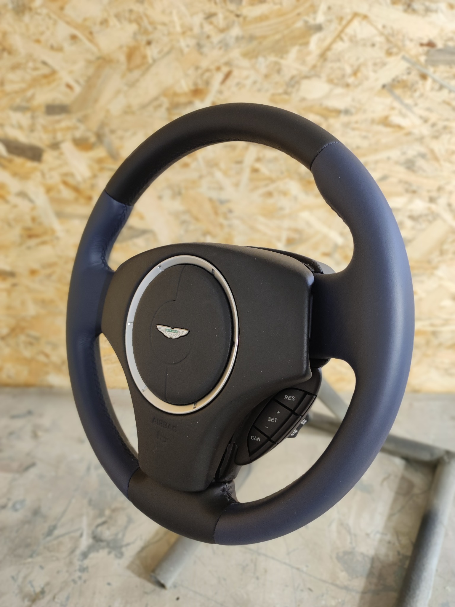 Odnowiona kierownica Aston Martin z czarną skórą na środku i nową granatową skórą na obręczy, widoczne przyciski sterowania i logo firmy.