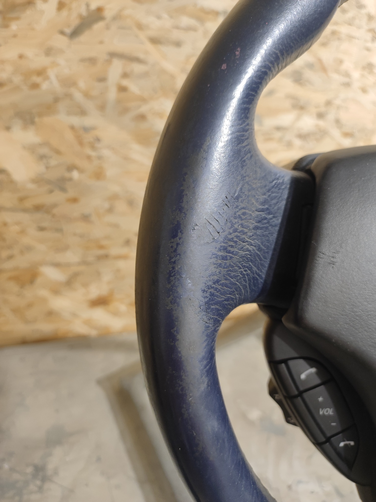 Zużyta, niebieska kierownica samochodowa z widocznymi śladami użytkowania i wytarciami na skórzanym obiciu, z przyciskami sterowania po prawej stronie, na tle płyty OSB.