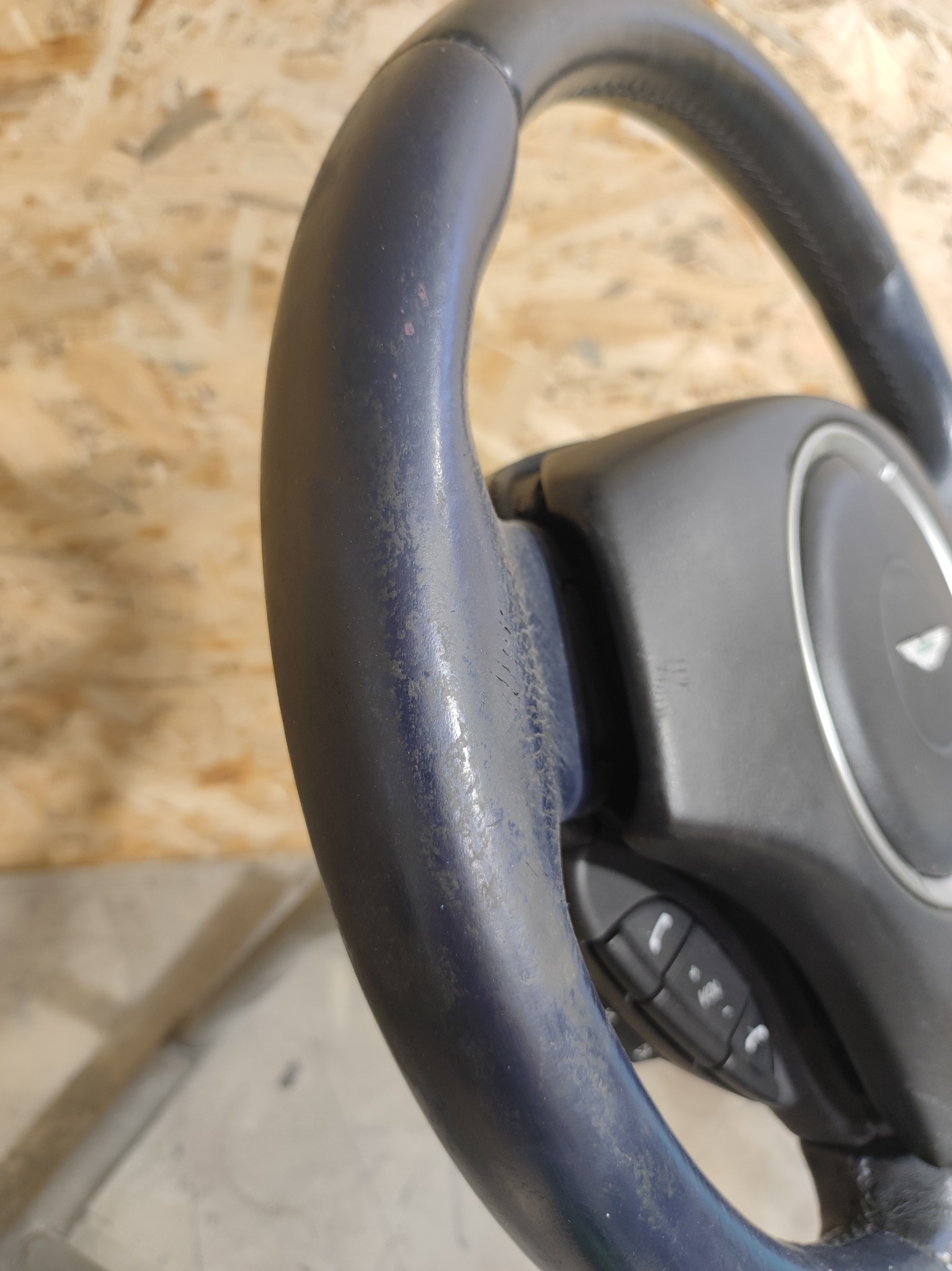 Zużyta, ciemna kierownica samochodowa z widocznymi śladami użytkowania i przyciskami funkcyjnymi, na tle drewnianej płyty.