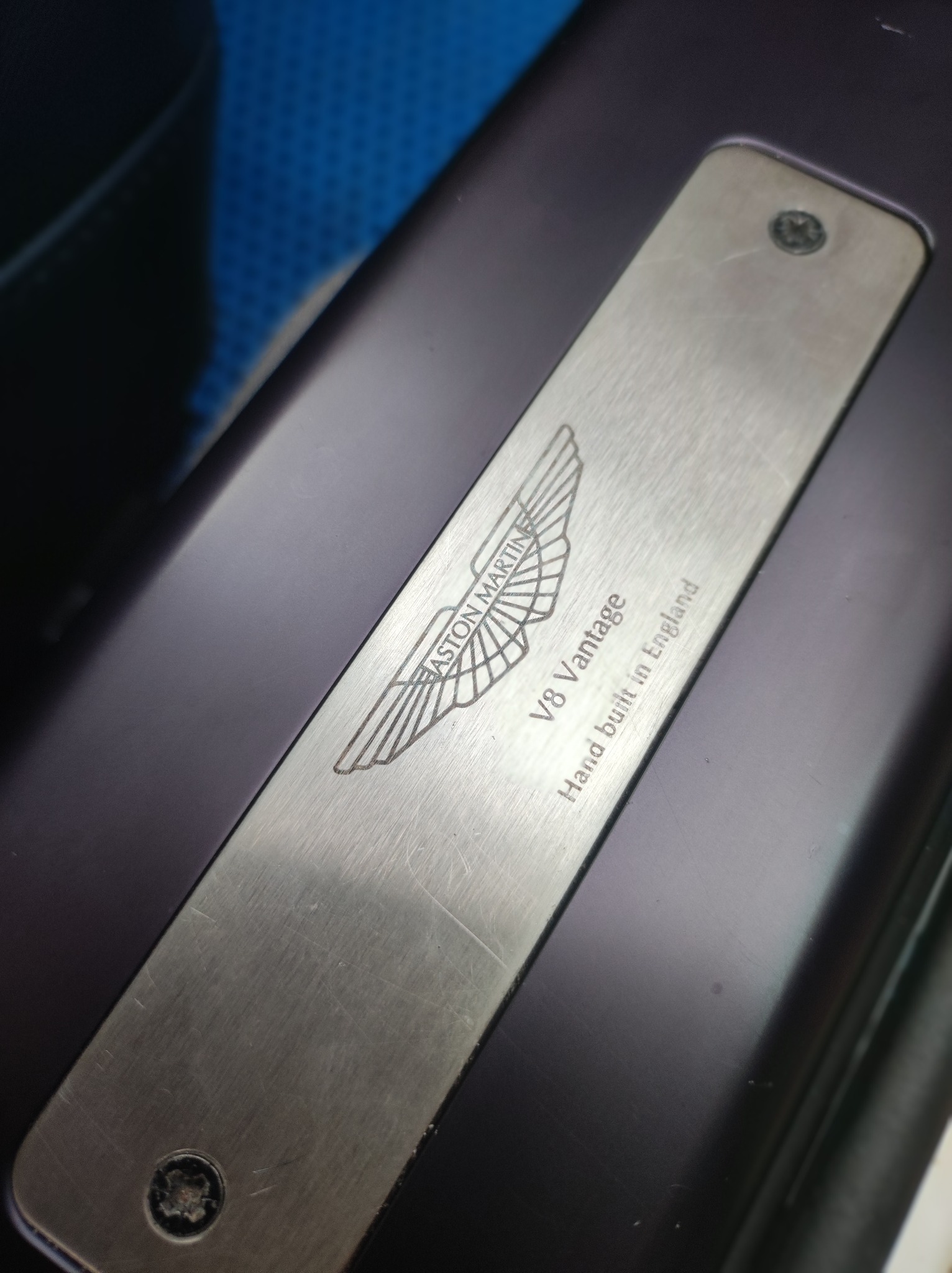 Metalowa tabliczka z wygrawerowanym logo Aston Martin V8 Vantage, przymocowana śrubami do ciemnofioletowej powierzchni.