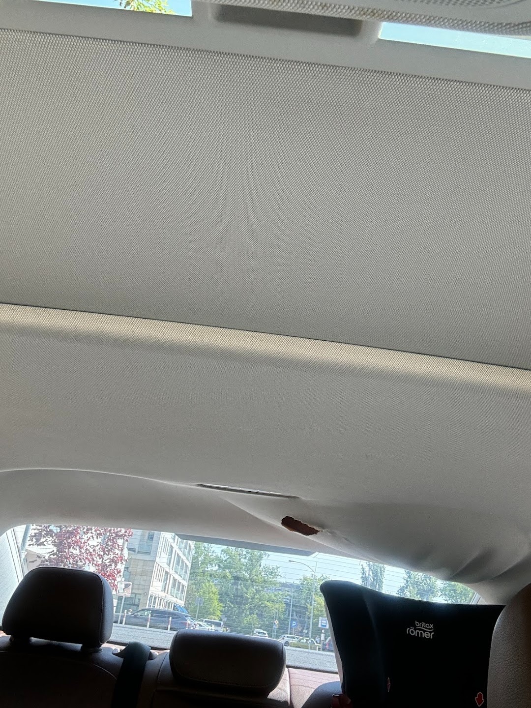 Uszkodzona podsufitka w samochodzie z widocznym ubytkiem materiału. Z tyłu widoczne zagłówki foteli i fotelik dziecięcy.