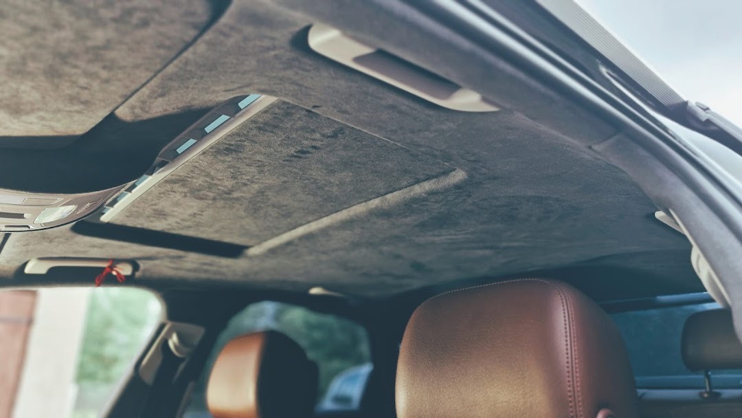 Wnętrze samochodu z nową, szarą podsufitką z alcantary i brązowymi skórzanymi siedzeniami, widoczne elementy oświetlenia i szyberdach.