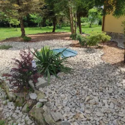 Ogród z wysypanym białym kamieniem, ozdobnymi krzewami, lampą ogrodową i zielonym włazem studzienki kanalizacyjnej.