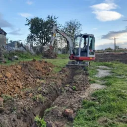 Mała koparka Takeuchi wykonuje wąski wykop w ziemi na działce, widoczne cegły i sterta ziemi obok, w tle zabudowania i drzewa pod błękitnym niebem.