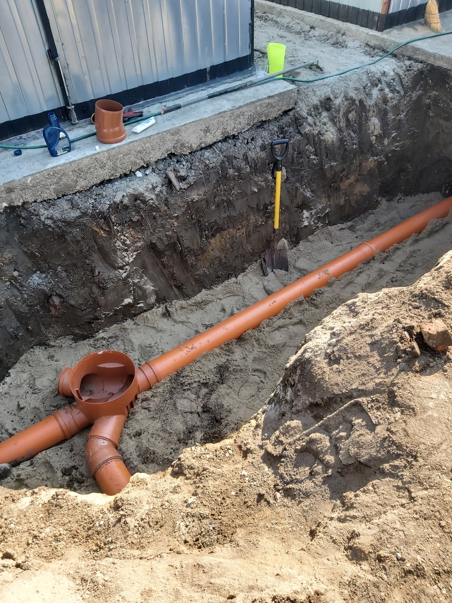 Pomarańczowe rury kanalizacyjne ułożone w wykopie wypełnionym piaskiem, z widoczną łopatą wbita w ścianę wykopu i narzędziami oraz pojemnikami na krawędzi wykopu.