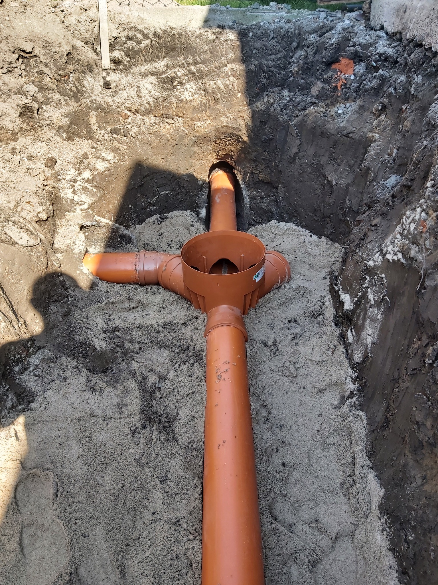 Pomarańczowe rury kanalizacyjne ułożone w wykopie wypełnionym piaskiem, widoczne w Pakosławicach. Ciemna ziemia po bokach wykopu, cień osoby w lewym dolnym rogu.