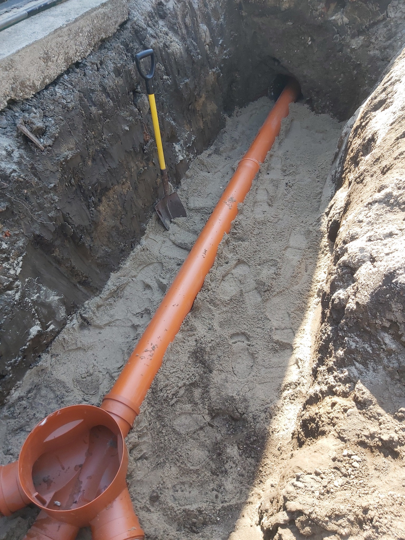 Pomarańczowa rura kanalizacyjna ułożona w wykopie wypełnionym piaskiem, obok stoi łopata z żółtym trzonkiem.
