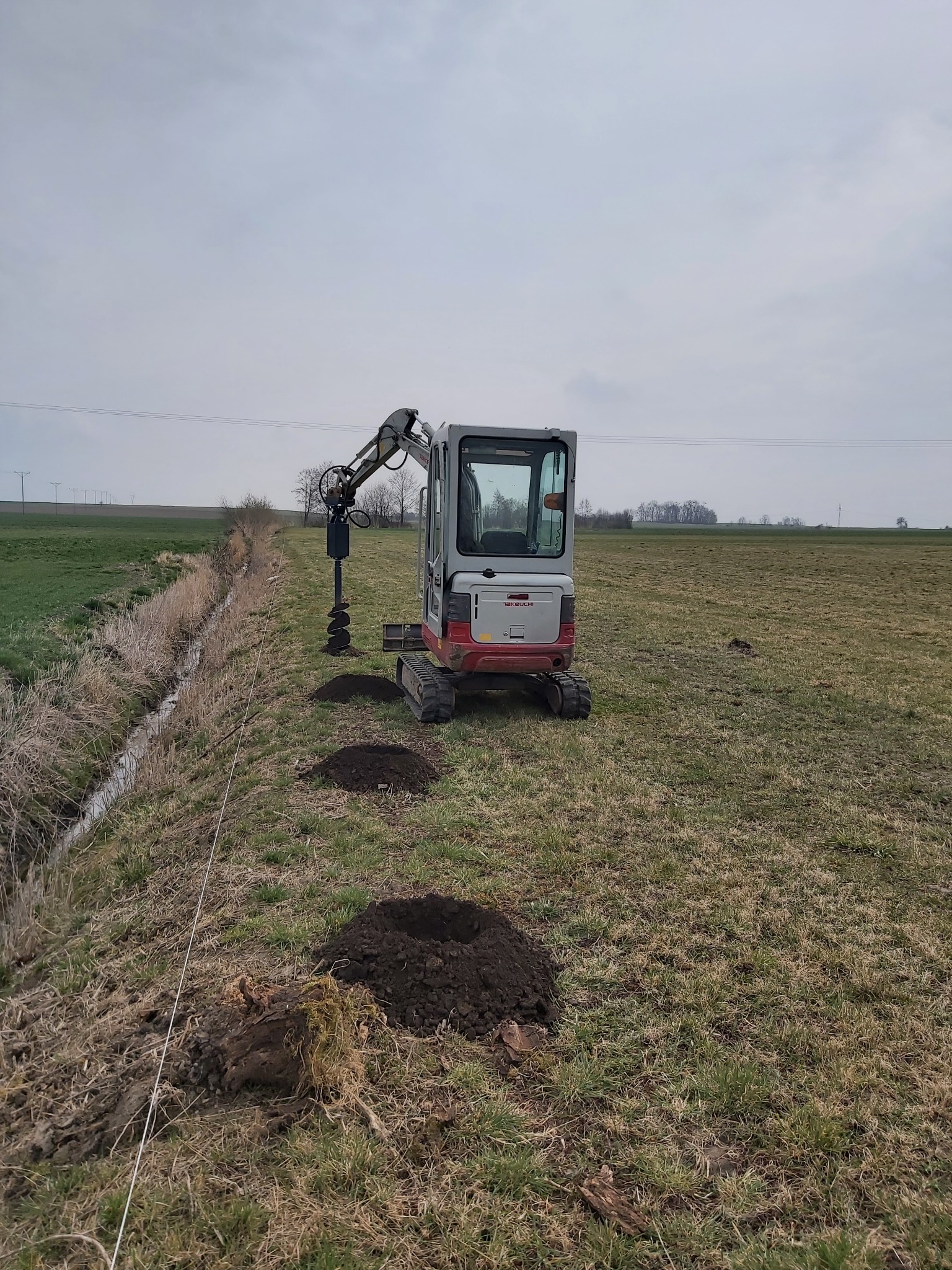 Mini koparka z wiertnicą hydrauliczną wykonuje otwory w ziemi wzdłuż rowu melioracyjnego na polu, widoczne usypane pryzmy ziemi.