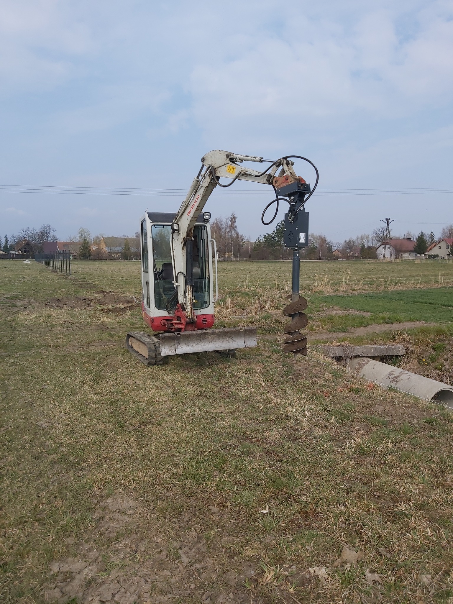 Mini koparka z wiertłem świdrowym w trakcie wiercenia otworu w ziemi na łące, widoczne rury betonowe w pobliżu.
