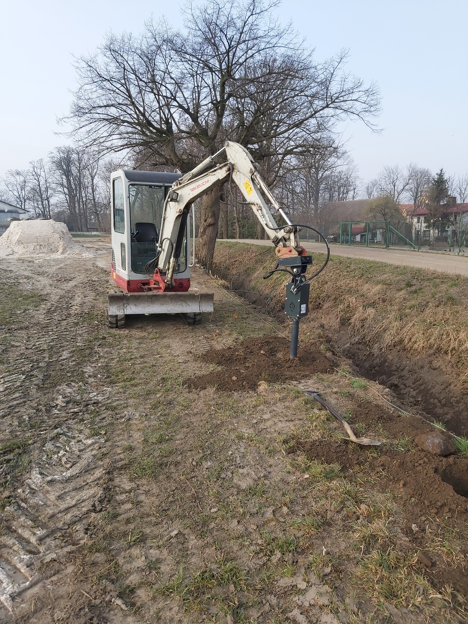 Mini koparka Takeuchi z wiertnicą hydrauliczną wiercąca otwór w ziemi wzdłuż wykopu, obok leży łopata, w tle sterta piasku i drzewa.