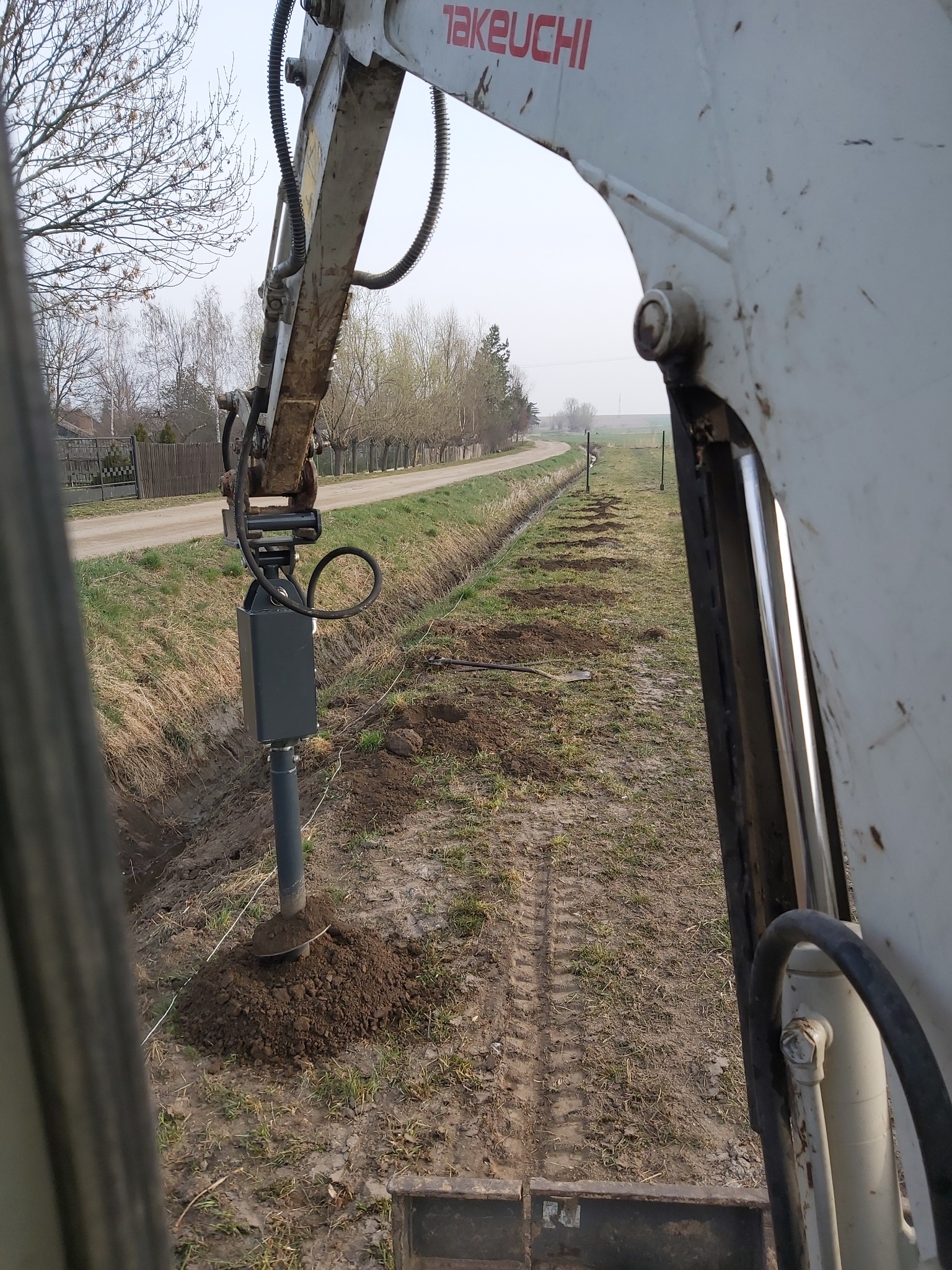 Maszyna Takeuchi w trakcie wbijania słupków w ziemię wzdłuż rowu, widok z kabiny operatora na rząd wykopanych dołków i krajobraz polny.