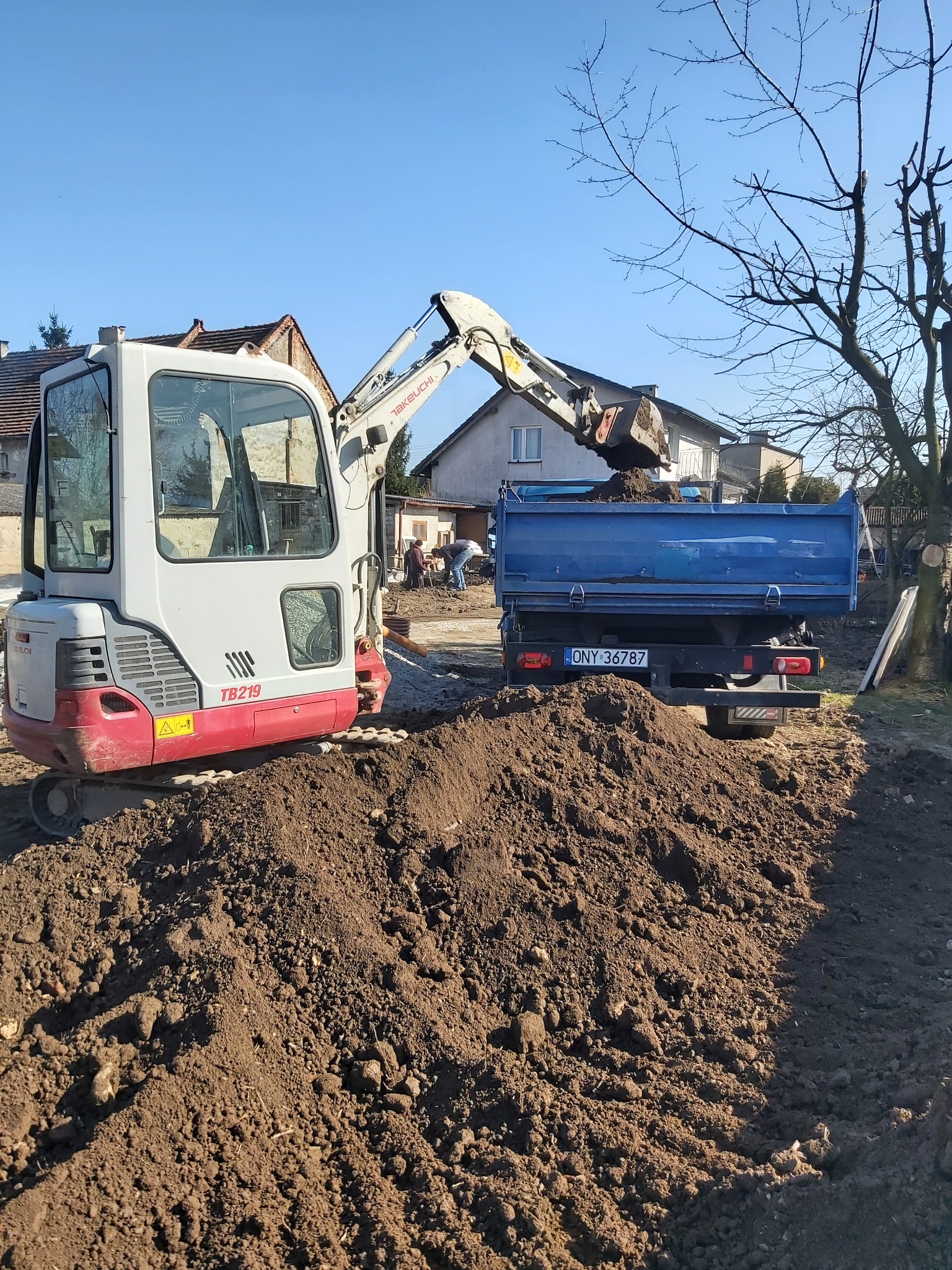 Kompaktowa koparka Takeuchi TB219 załadowuje ziemię do niebieskiej wywrotki na tle zabudowy jednorodzinnej w słoneczny dzień.