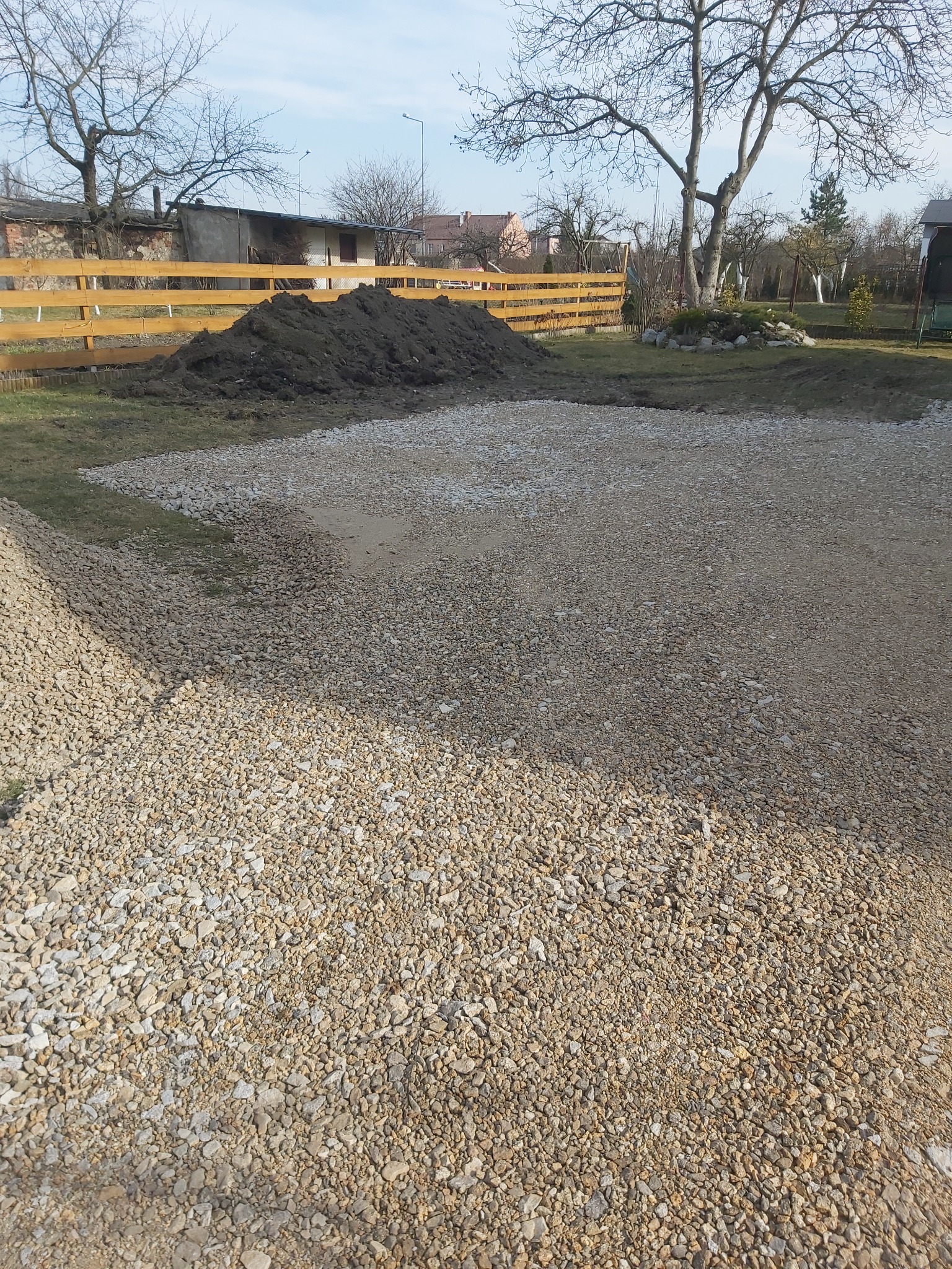 Teren przygotowany pod budowę, wysypany żwirem, z widoczną pryzmą ziemi w tle i drewnianym ogrodzeniem.