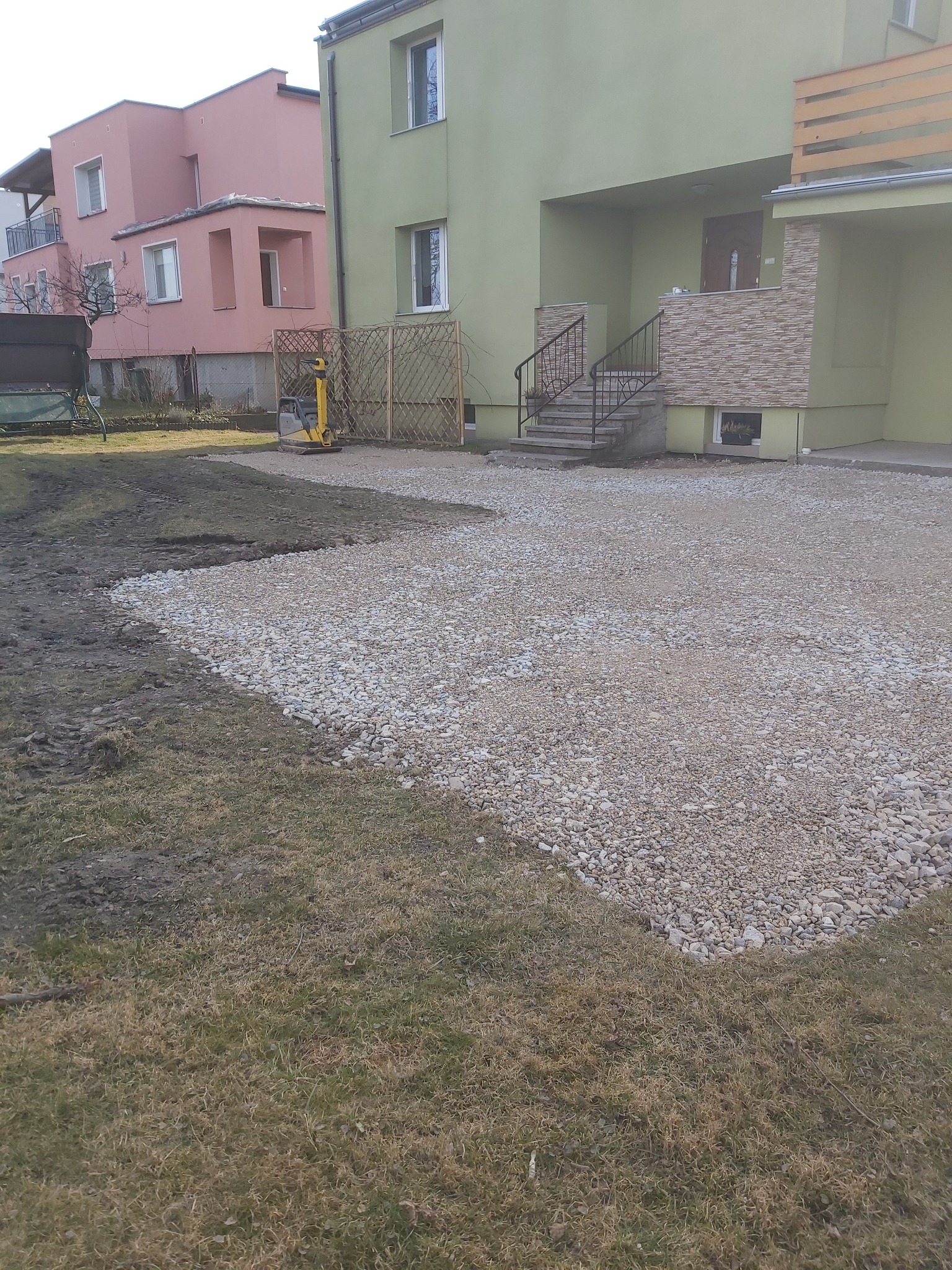 Przygotowanie terenu pod budowę: wyrównany teren z warstwą kamieni, zagęszczarka w tle, trawnik i budynki mieszkalne.