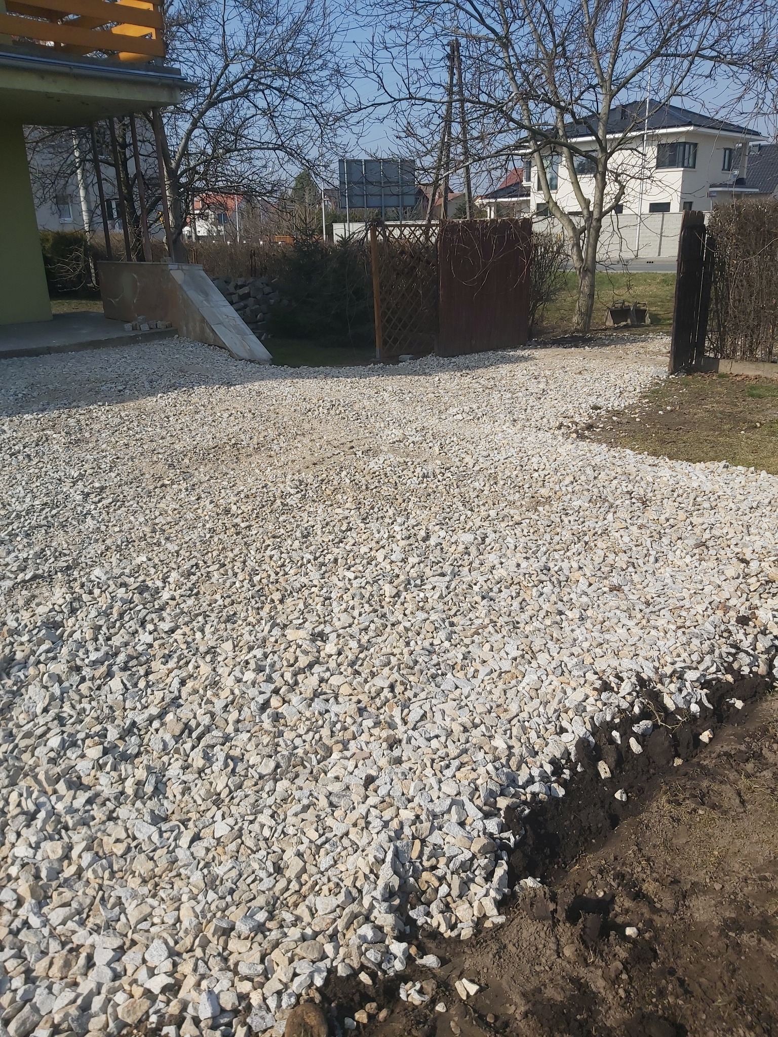 Ułożona warstwa białego kruszywa na podwórku z widocznym obrzeżem z ziemi oraz budynkami w tle.