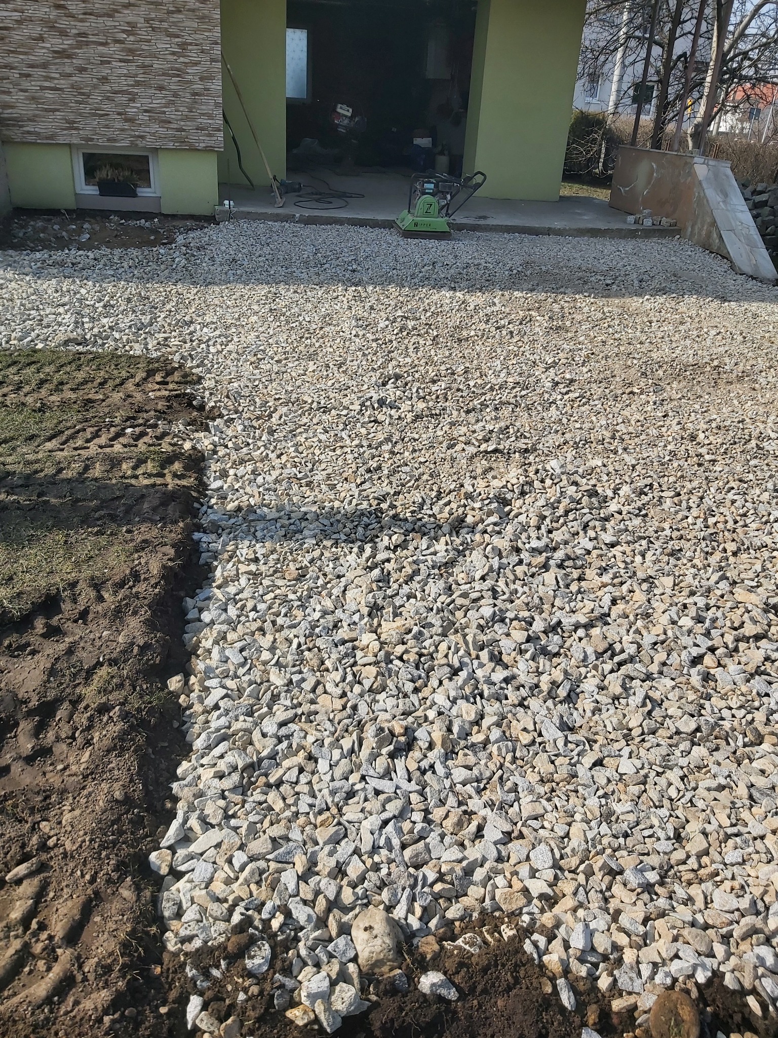 Przygotowanie terenu: wyrównany grunt z widoczną warstwą jasnego żwiru, obok fragment wykopanej ziemi i trawy, w tle budynek gospodarczy z zielonymi ścianami i zagęszczarka wibracyjna.