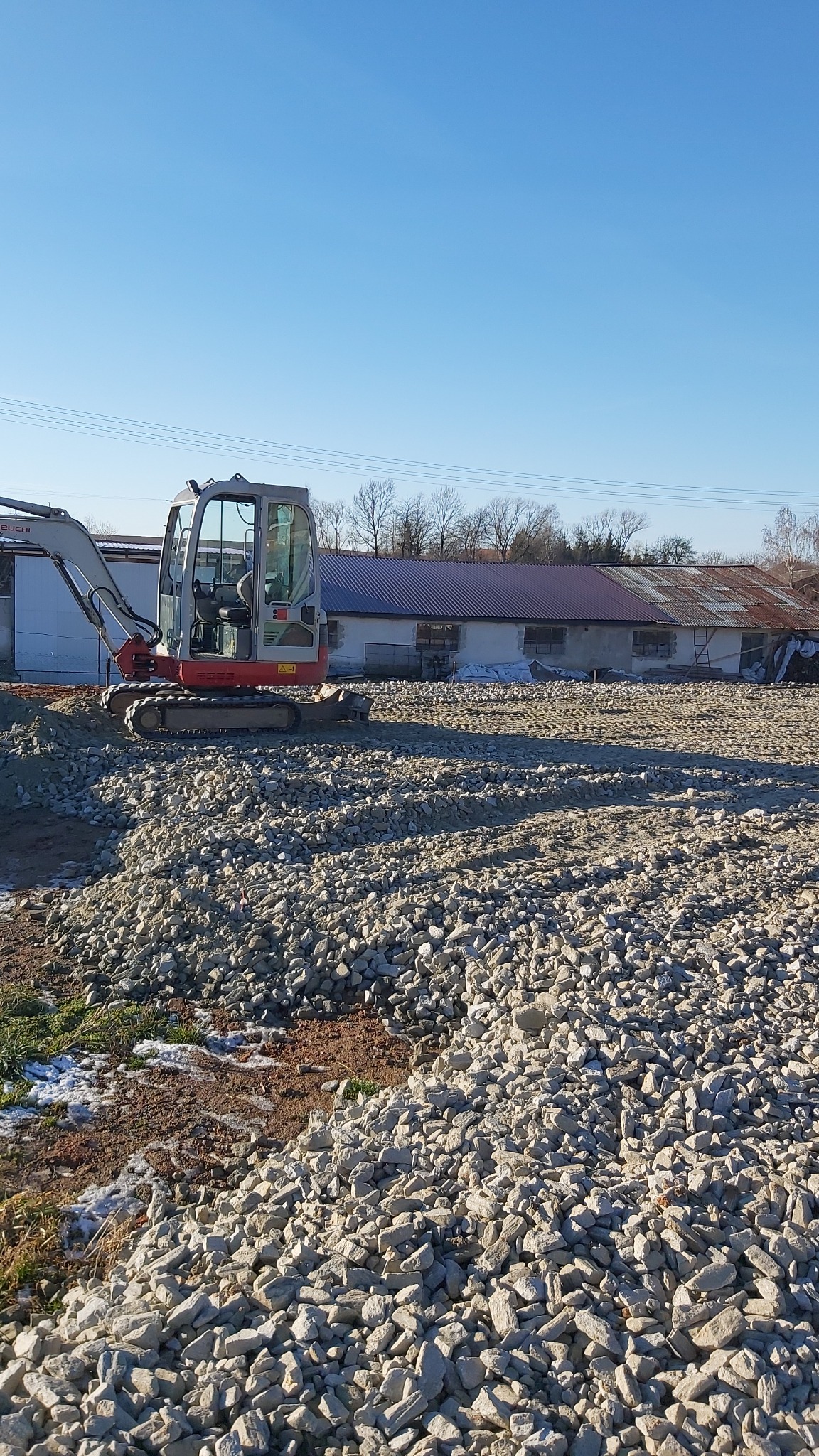Mini koparka Takeuchi stojąca na wyrównanym terenie wysypanym gruzem, w tle budynek gospodarczy z blachodachówką i błękitne niebo.