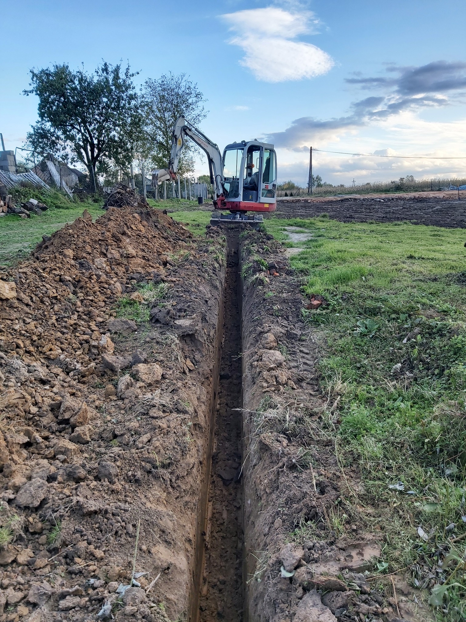 Wąski wykop liniowy w gruncie, z mini-koparką na końcu wykopu, w tle drzewa i zabudowania gospodarcze.