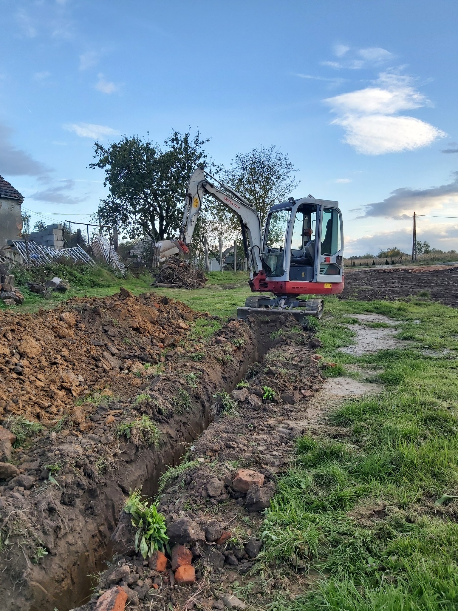 Mała koparka Takeuchi wykonuje wąski wykop w ziemi na działce, widoczne cegły i sterta ziemi obok, w tle zabudowania i drzewa pod błękitnym niebem.