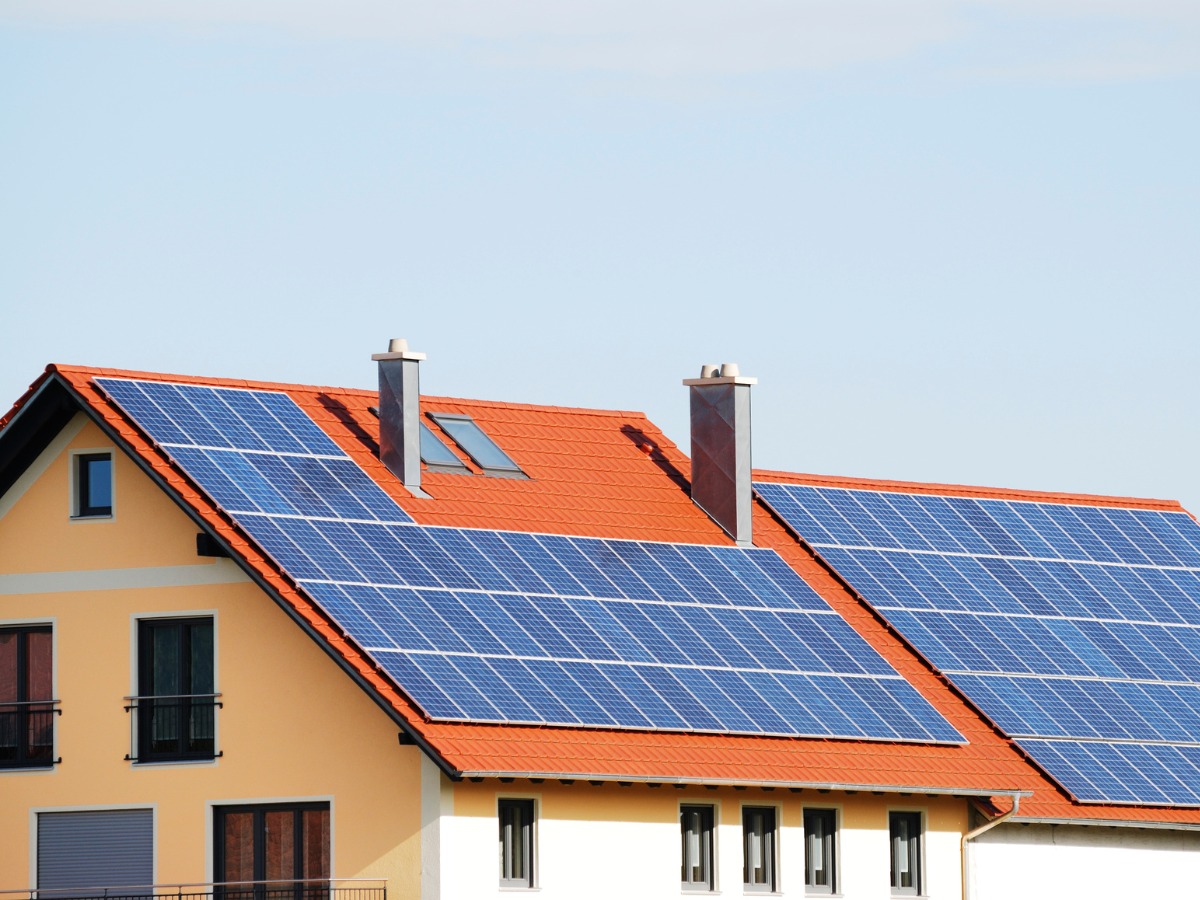 Żółty dom z czerwonym dachem, na którym zamontowane są panele fotowoltaiczne. Widoczne okna dachowe i komin.
