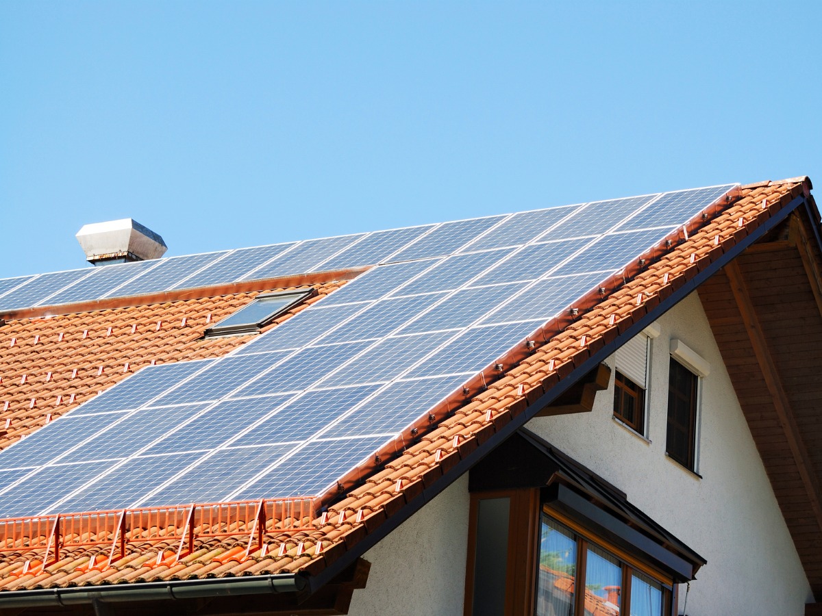 Instalacja fotowoltaiczna na dachu domu jednorodzinnego z czerwoną dachówką. Widoczne okno dachowe i komin. Błękitne niebo w tle.