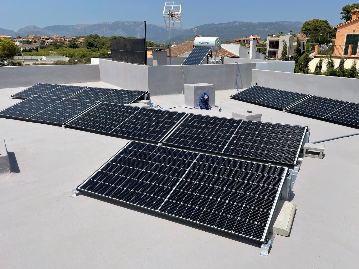 Instalacja fotowoltaiczna na płaskim dachu z widokiem na góry. Panele w czarnych ramach, szare podłoże dachu. Widoczny zasobnik na wodę i antena.