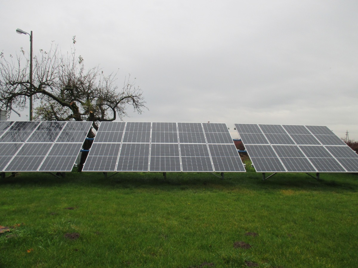 Instalacja fotowoltaiczna na trawie pod pochmurnym niebem, z widocznym drzewem w tle. Panele ustawione w trzech rzędach, równomiernie rozmieszczone na zielonym terenie.