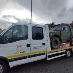 Biała laweta z logo firmy 'HELP MODE' przewozi terenowy samochód Suzuki Samurai w kolorze khaki, zabezpieczony pomarańczowymi pasami transportowymi.