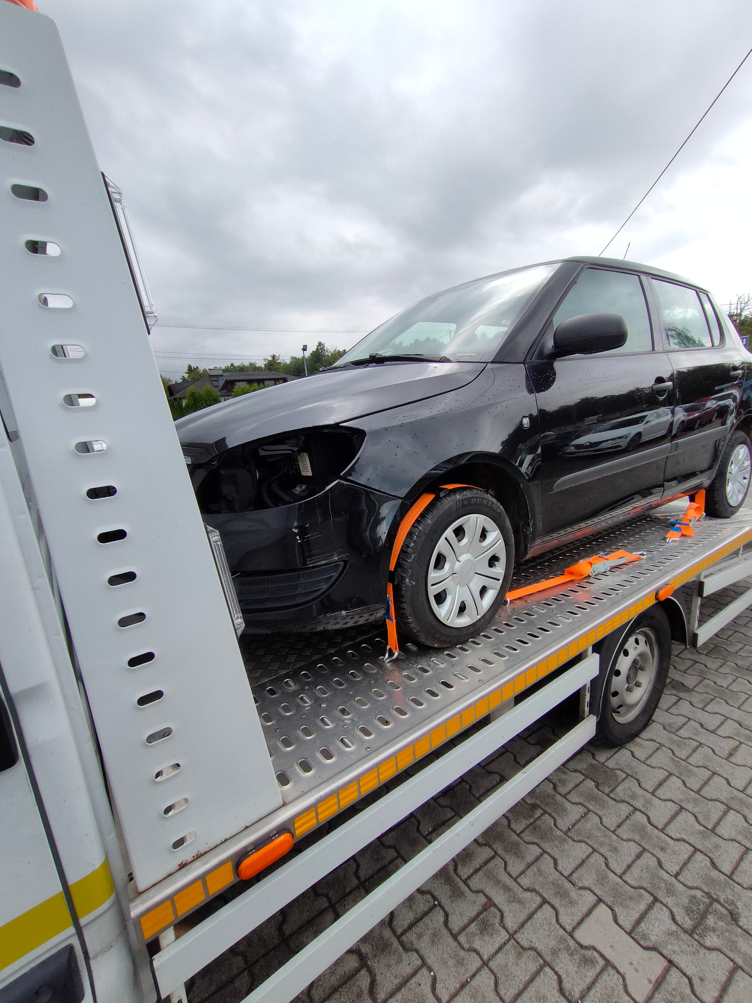 Czarny uszkodzony samochód osobowy, zabezpieczony pomarańczowymi pasami, transportowany na lawecie na tle pochmurnego nieba.
