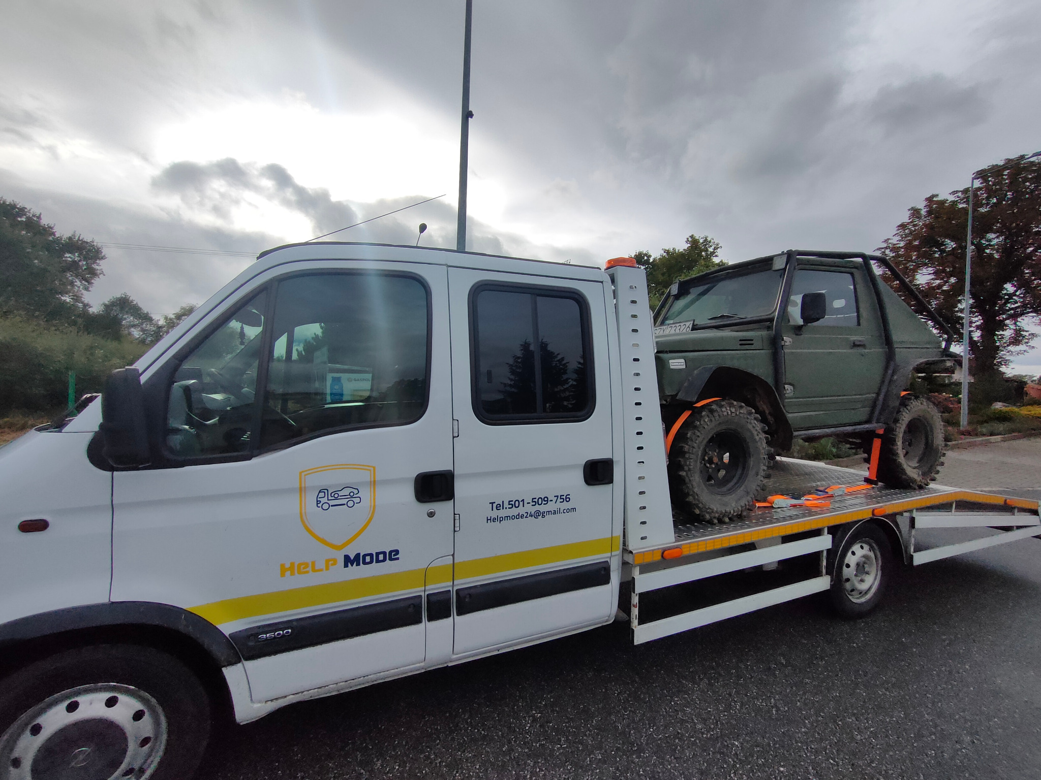 Biała laweta z logo firmy 'HELP MODE' przewozi terenowy samochód Suzuki Samurai w kolorze khaki, zabezpieczony pomarańczowymi pasami transportowymi.