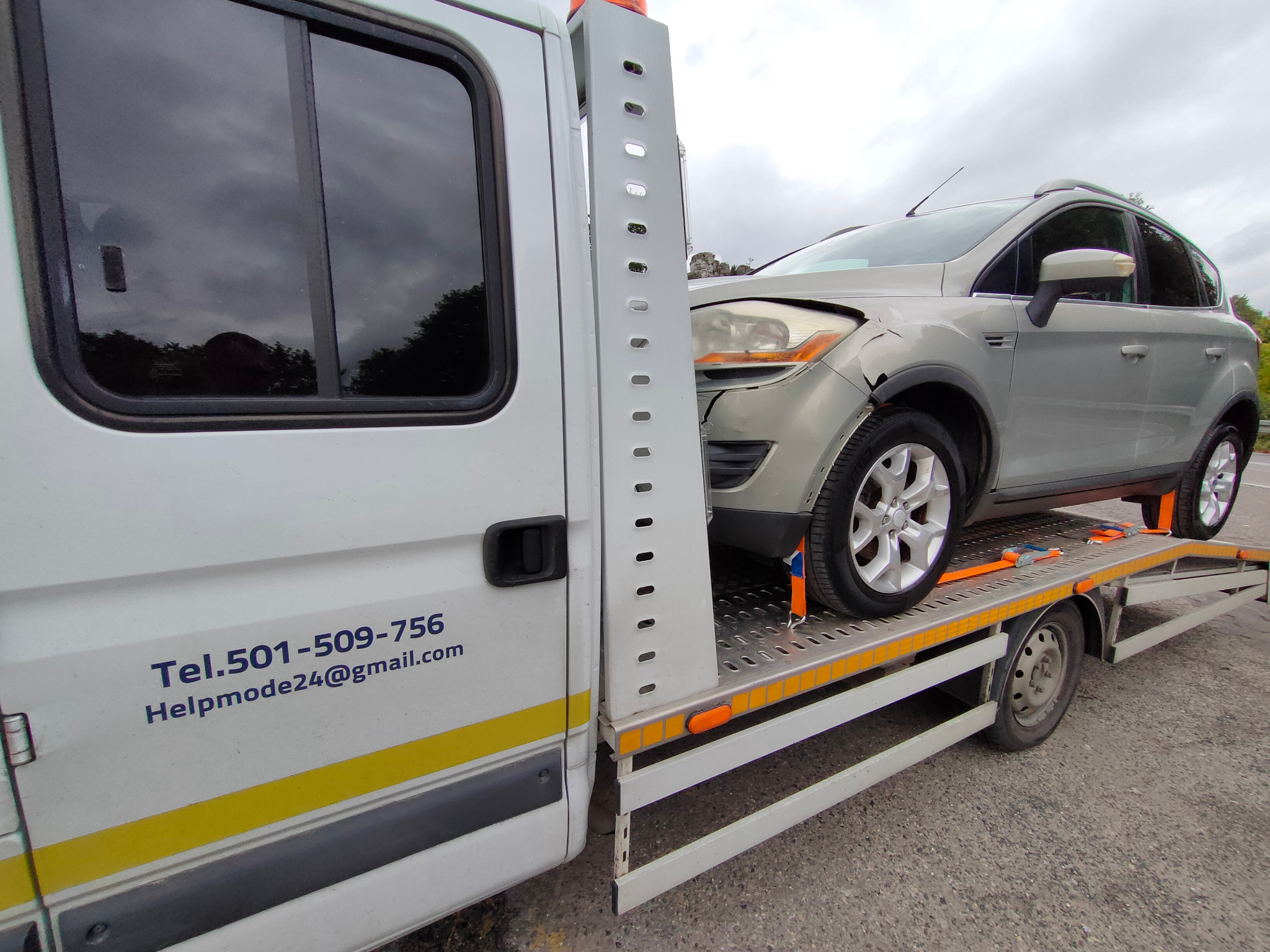 Uszkodzony srebrny samochód osobowy, zabezpieczony pomarańczowymi pasami, przewożony na platformie białej ciężarówki z widocznym numerem telefonu i adresem e-mail.