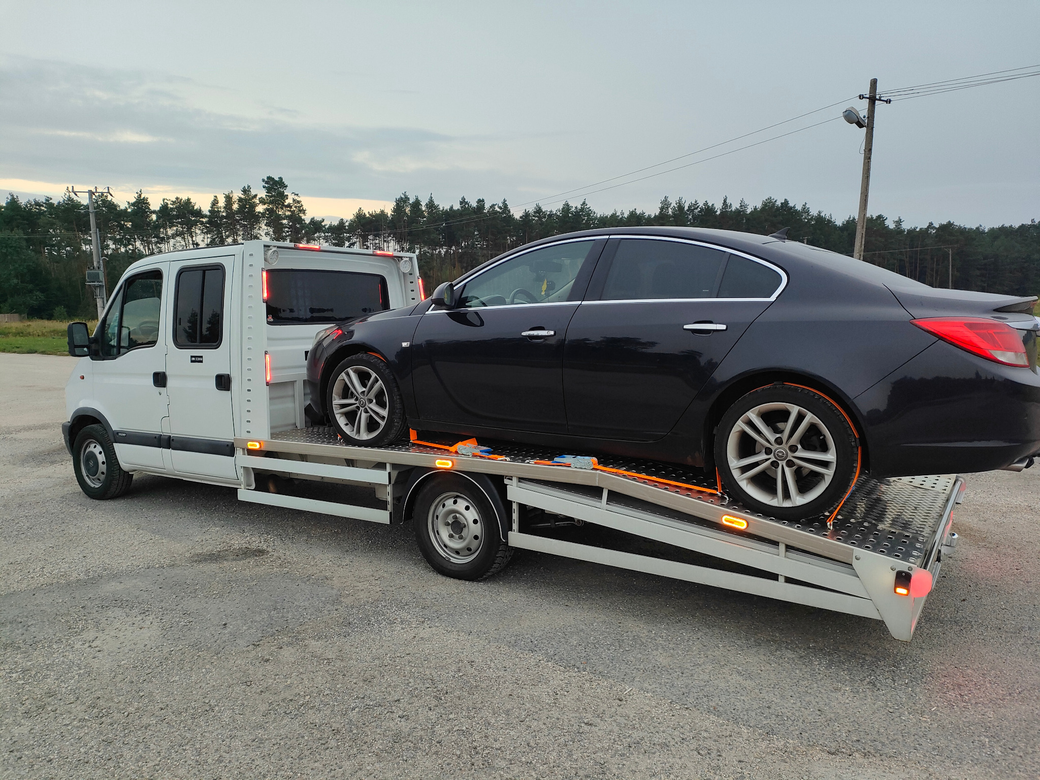 Czarny samochód osobowy marki Opel Insignia zabezpieczony pomarańczowymi pasami transportowymi na platformie białej lawety w trakcie transportu.