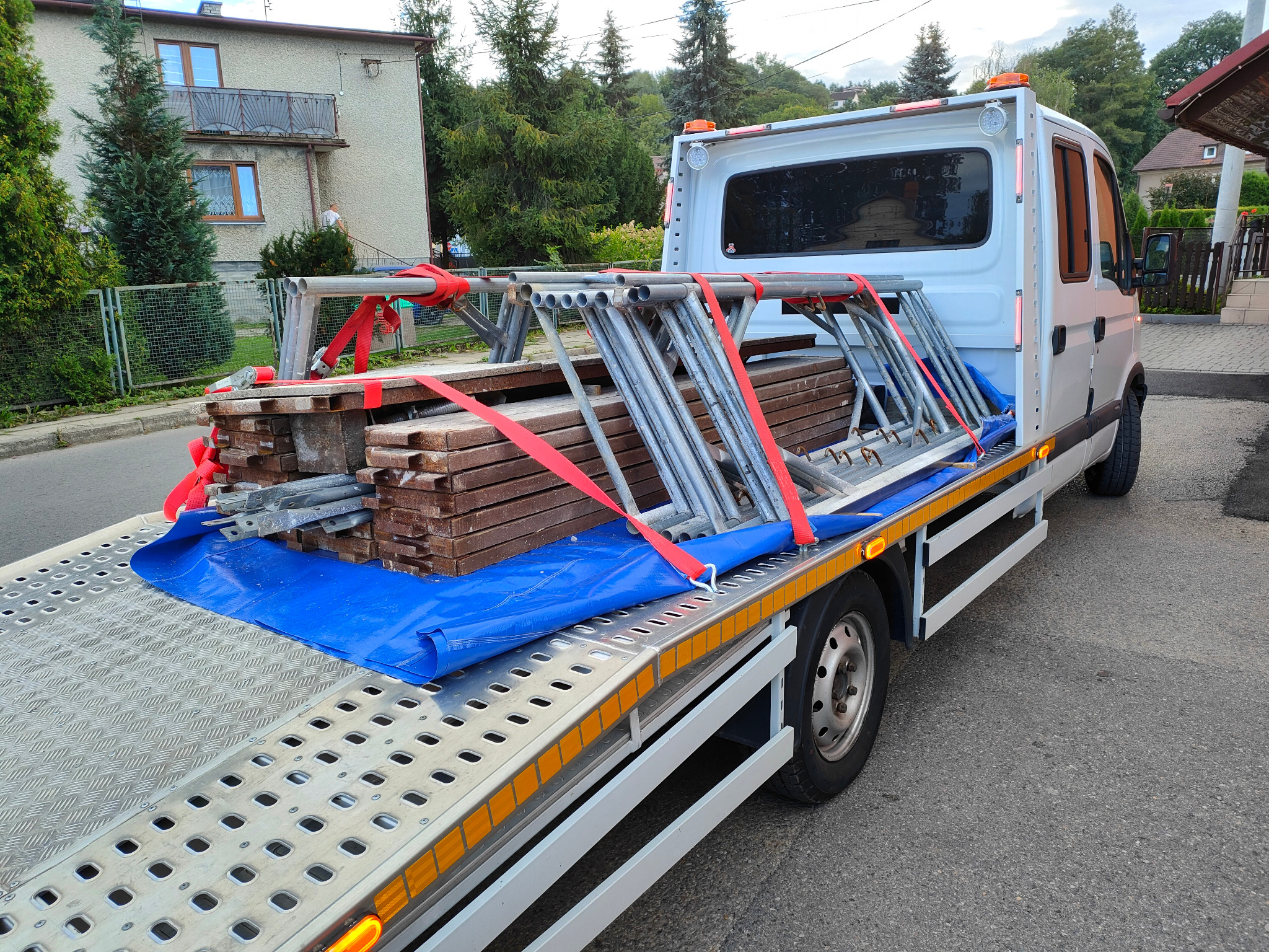 Biała platforma transportowa przewozi zabezpieczony czerwonymi pasami ładunek desek i metalowych rusztowań na tle miejskiej zabudowy i zieleni.