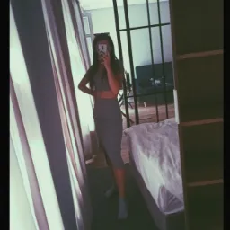 Kobieta w szarym komplecie pozuje do selfie w lustrze w pokoju z zasłoniętym oknem i łóżkiem