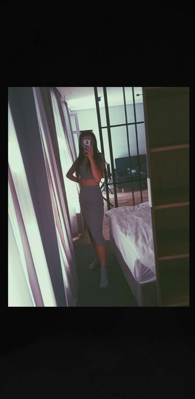 Kobieta w szarym komplecie pozuje do selfie w lustrze w pokoju z zasłoniętym oknem i łóżkiem