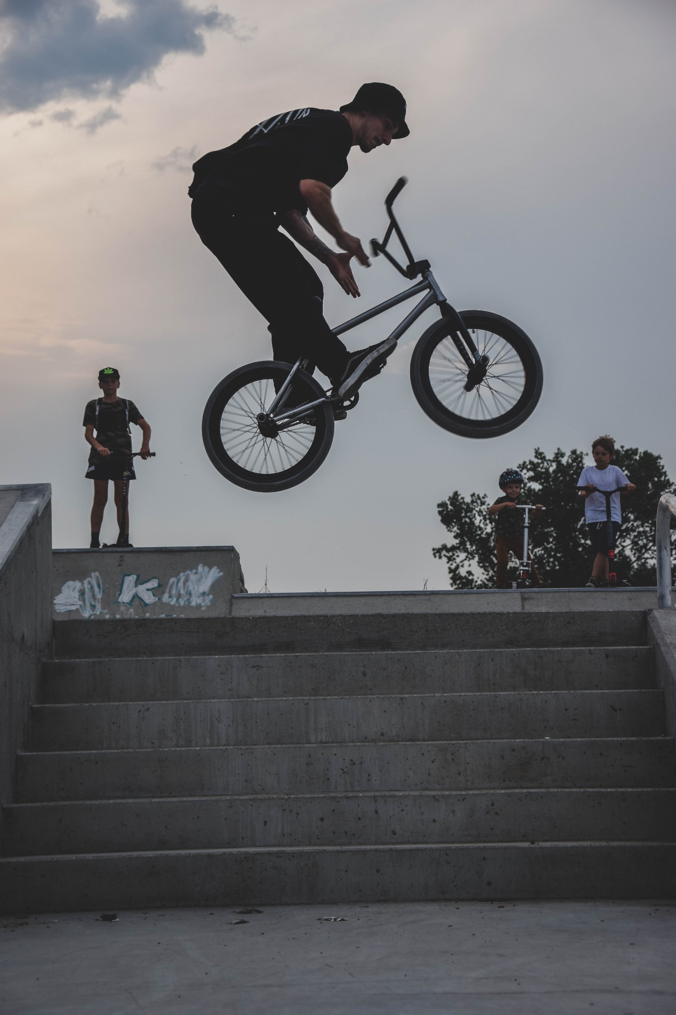 Mężczyzna na rowerze BMX wykonuje trik w powietrzu w skateparku, w tle dzieci na hulajnogach i betonowe schody.