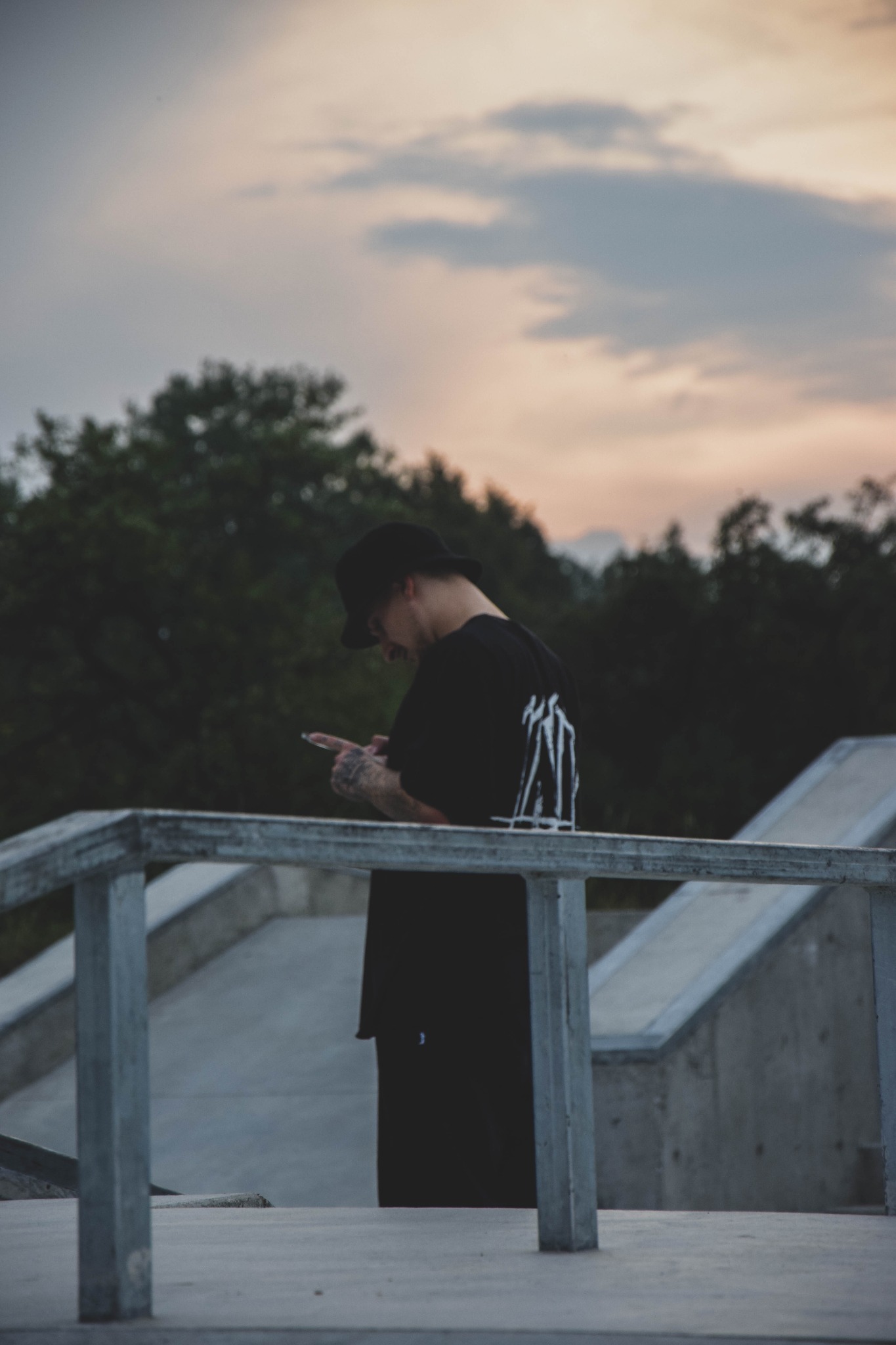 Mężczyzna w czarnym ubraniu i kapeluszu, z tatuażami na rękach, korzysta z telefonu w skateparku o zmierzchu, opierając się o metalową barierkę.